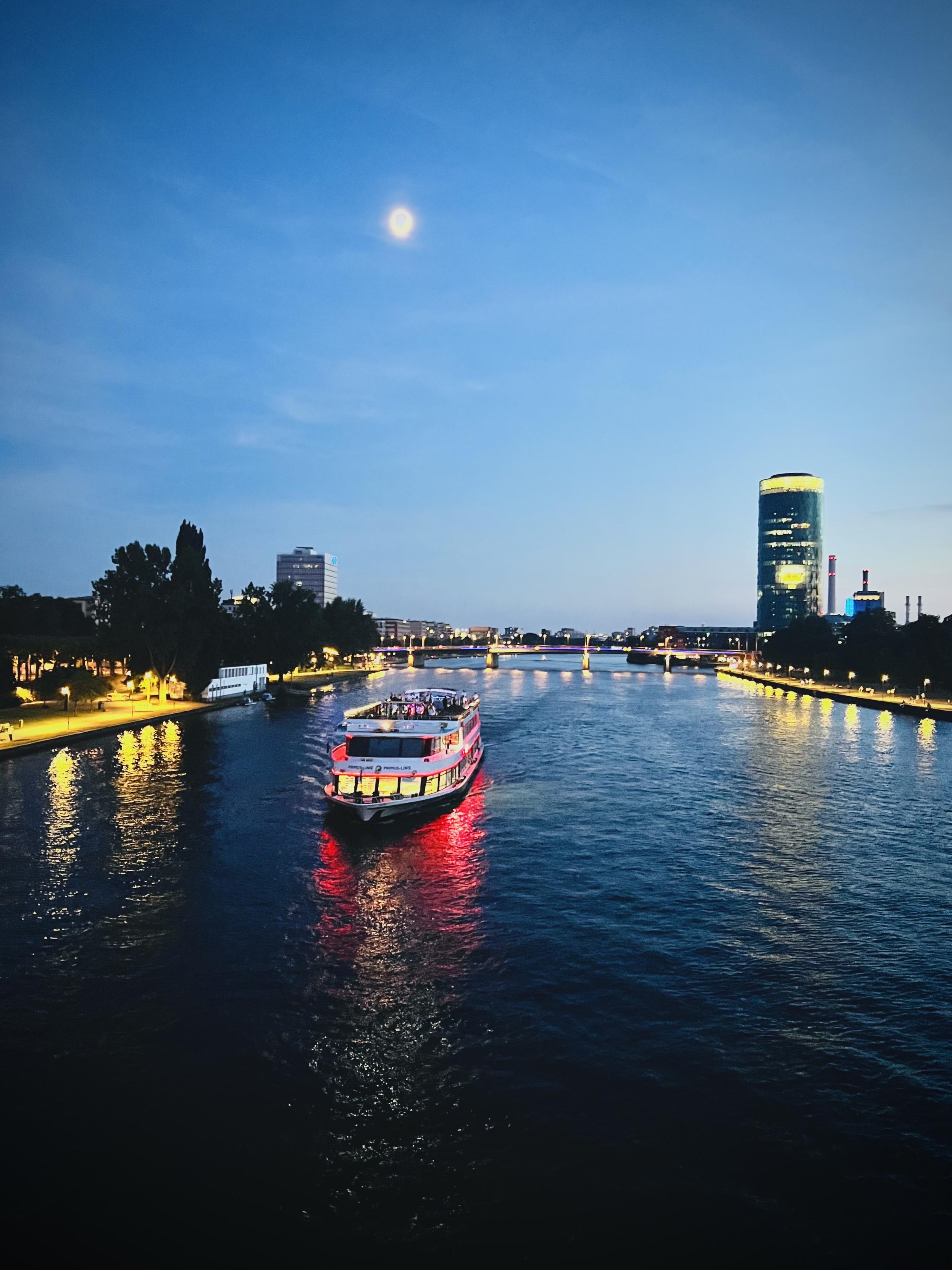 An illuminated passenger boat cruises down the center of a wide river at twilight, casting a red and golden glow on the water. A tall, cylindrical glass skyscraper stands lit on the right bank under a deep blue sky where a bright moon shines, while streetlights along the shores and distant bridges reflect across the dark river surface.