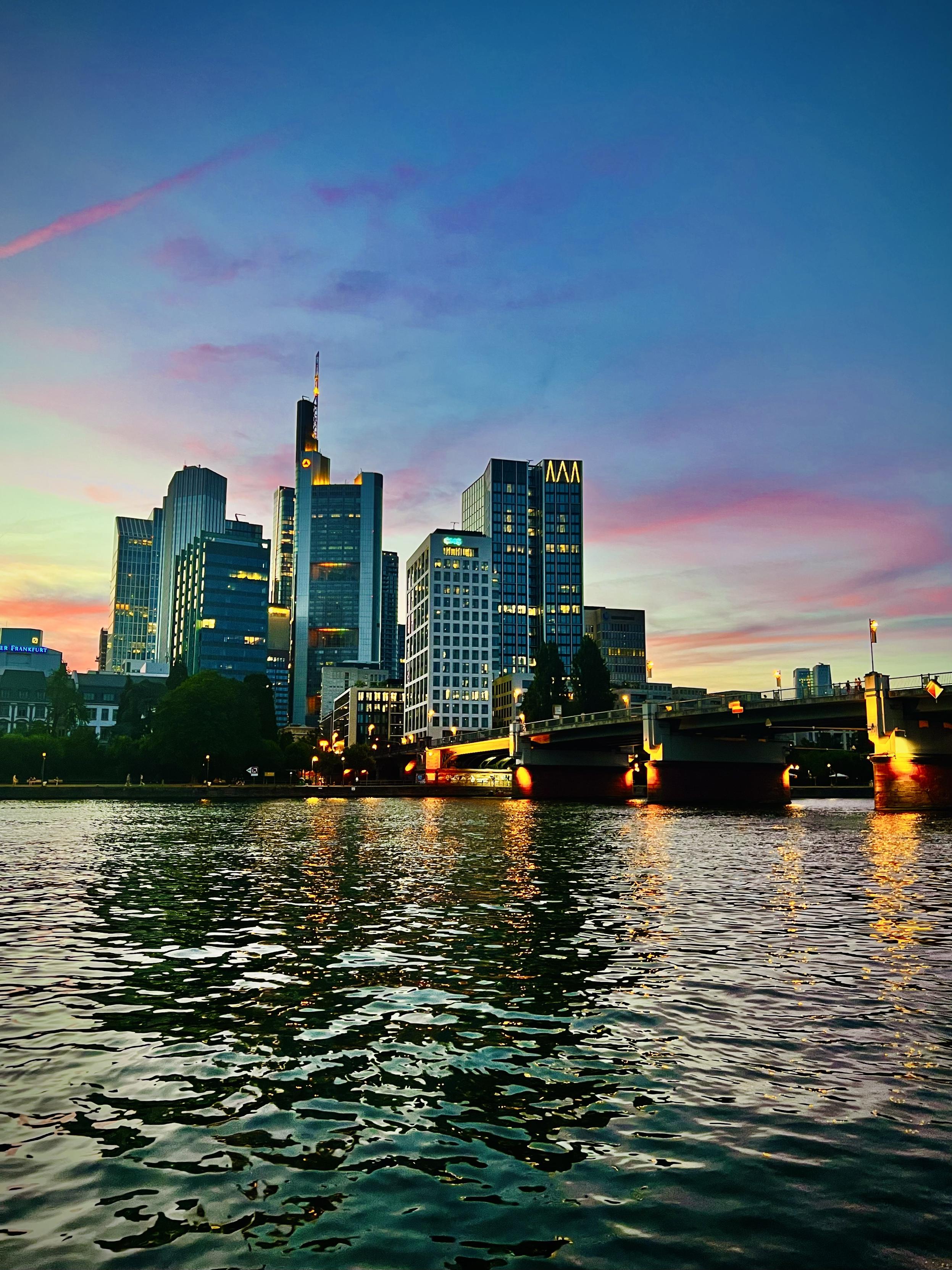 A vibrant sunset sky with streaks of pink, orange, and deep blue hangs over a modern city skyline. The high-rise skyscrapers are lit from within, standing behind a concrete bridge that spans a wide river. Warm yellow lights from the bridge and buildings reflect across the gently rippling surface of the dark water in the foreground.