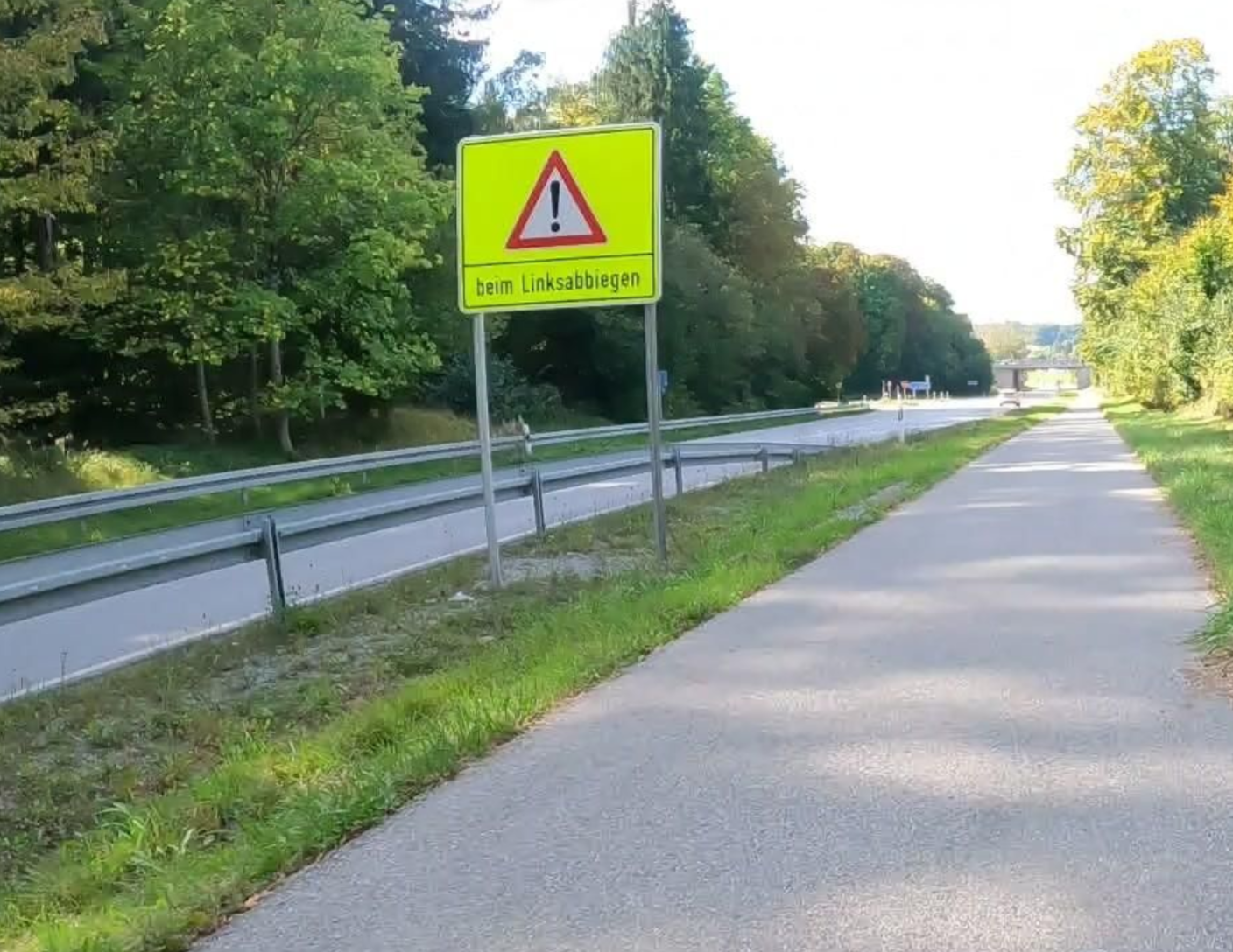 Blick auf einen Radweg mit Fahrbahn daneben. Neben der Fahrbahn steht ein gelbes Schild, das vor einer gefährlichen Stelle warnen soll. Darauf ist ein Achtung-Symbol und die Aufschrift "beim Linksabbiegen".