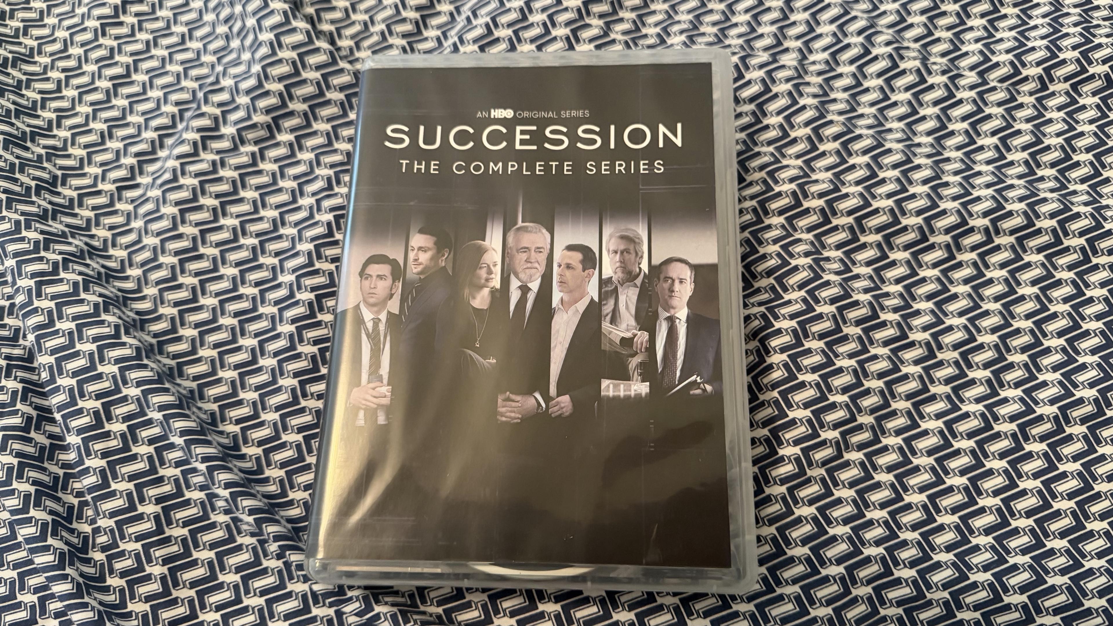 The image shows the DVD case for "Succession: The Complete Series," an HBO original series. The cover features several characters in professional attire, posing against a dark background. The case is placed on a patterned blue and white fabric.
