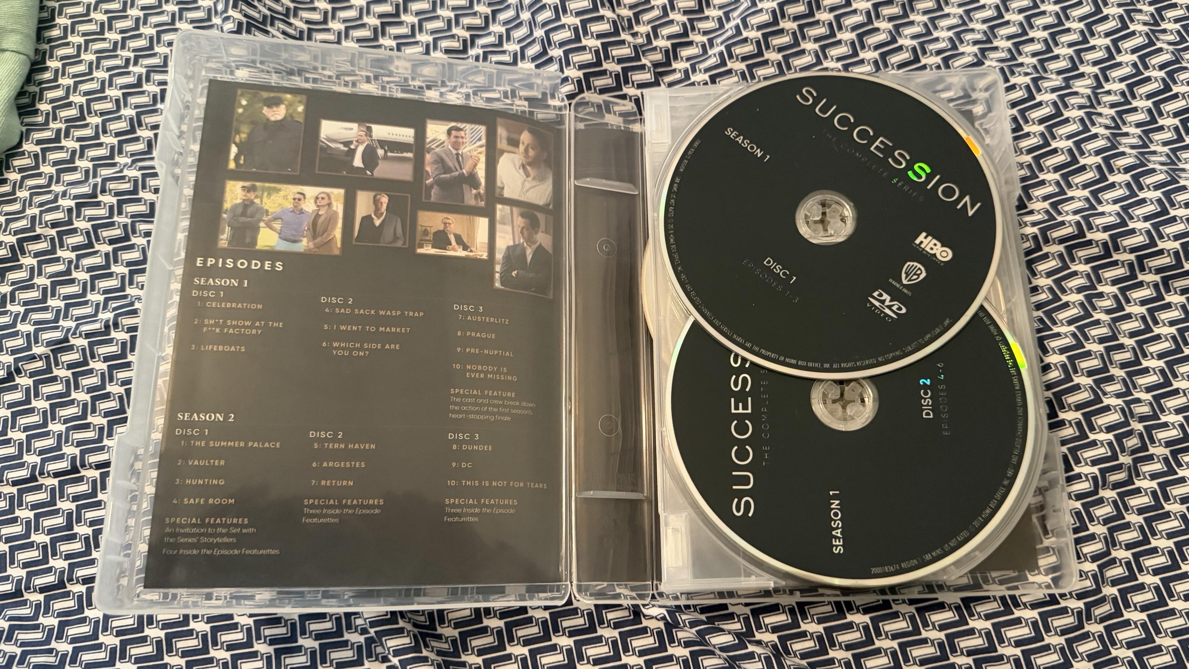 The image shows a DVD case for the TV series "Succession." It contains two discs labeled for Season 1, with detailed episode listings for both Season 1 and Season 2 printed inside the case. A patterned background is visible beneath the case