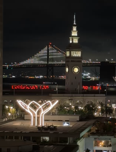 video of the San Francisco ferry building clock tower with colored lights rotating, and in the background of the San Francisco Bay Bridge with the white light display.