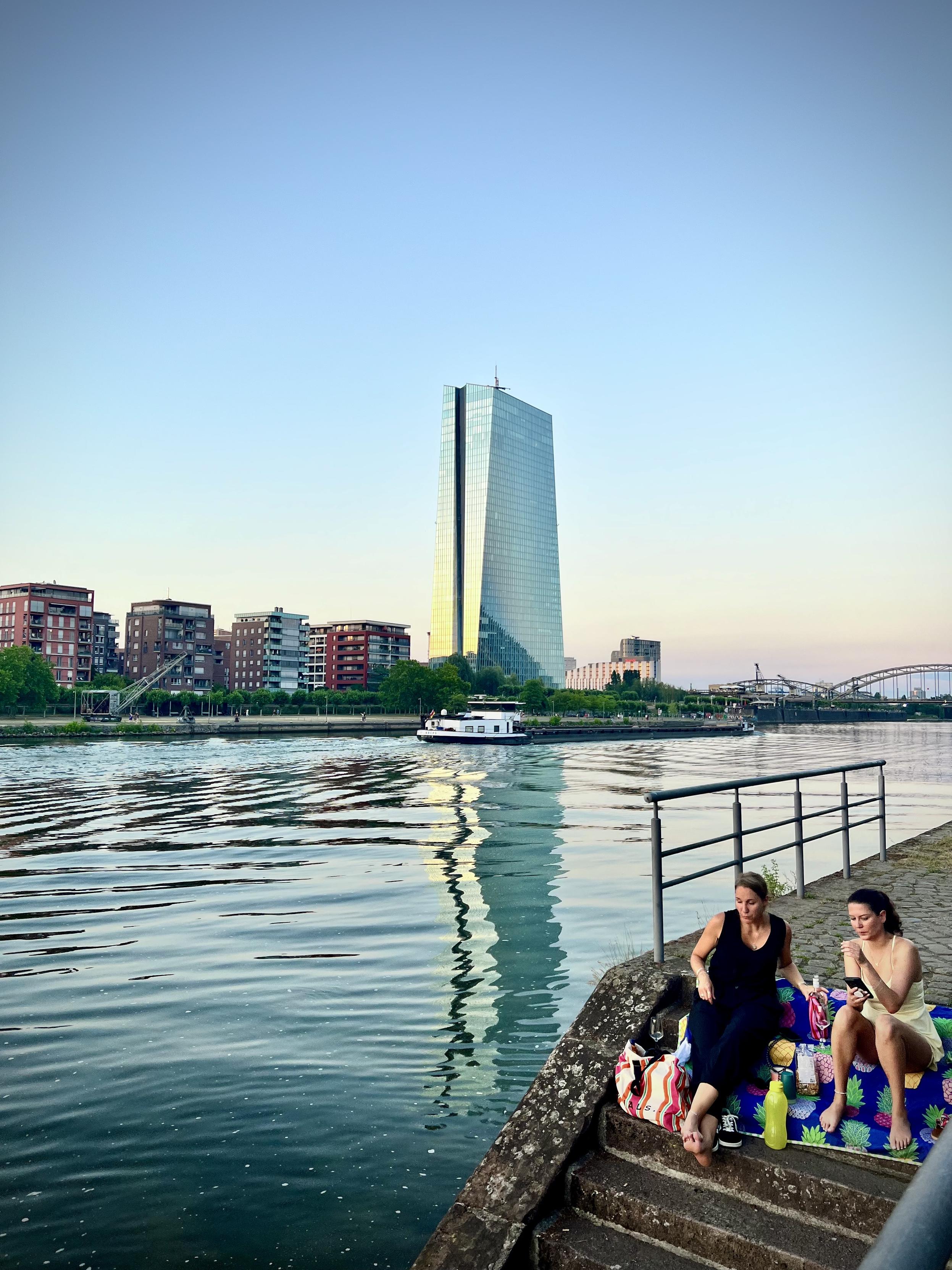 A wide river flows under a clear sky during the golden hour, with a prominent modern glass skyscraper on the opposite bank reflecting the warm sunlight and casting a long, wavy reflection in the water. A cargo barge travels down the river past mid-rise residential buildings. In the bottom-right foreground, two women sit on a colorful, pineapple-patterned blanket spread across stone steps next to a metal railing, relaxing by the waterfront.