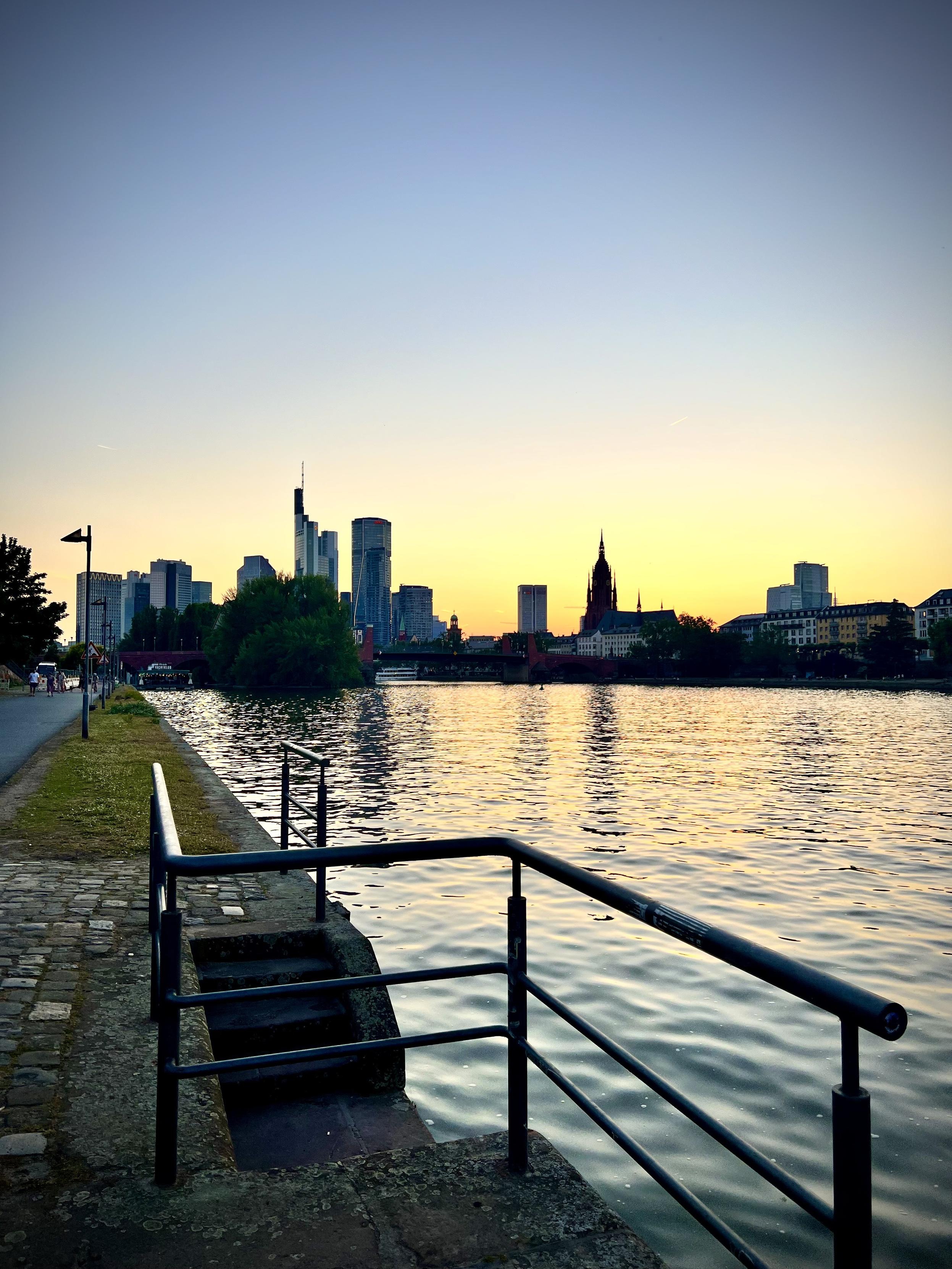A city skyline featuring a mix of modern skyscrapers and a historic gothic cathedral spire is silhouetted against a vibrant orange and yellow sunset. In the foreground, a stone riverbank with a cobblestone path and a black metal railing overlooks a wide river, which ripples with bright golden reflections from the evening sky.