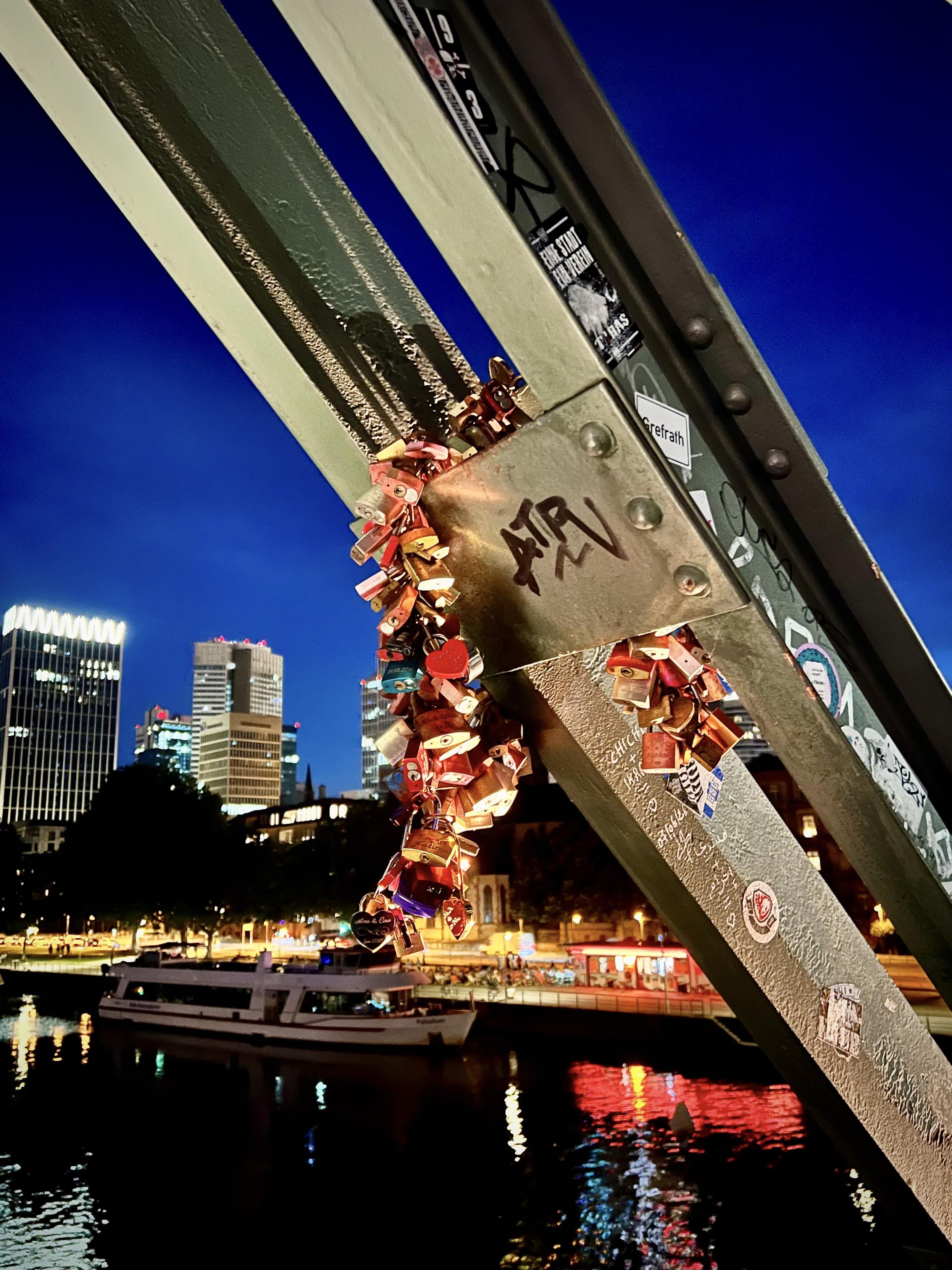 A close-up shot focuses on a cluster of colorful love padlocks attached to a metal bridge girder covered in stickers and graffiti. In the background, a tour boat sits on a dark river with vibrant light reflections, framed by a lit-up city skyline under a deep blue twilight sky.