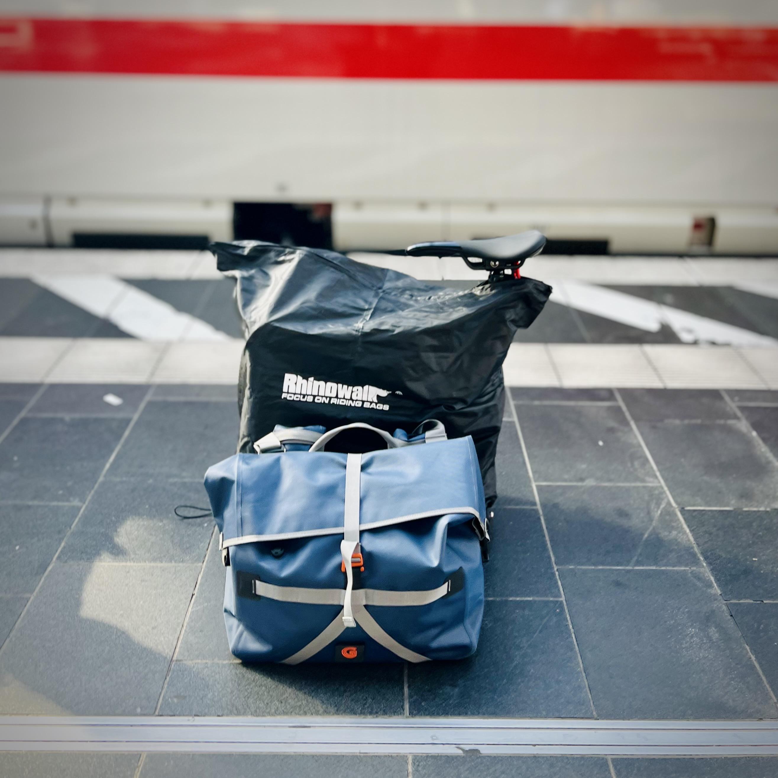 A blue, waterproof roll-top travel bag with grey straps sits on a tiled train platform in front of a larger, black Rhinowalk bike travel bag. My Brompton saddle protrudes from the top of the black bag, and a white train with a distinctive red stripe is softly blurred in the background.
