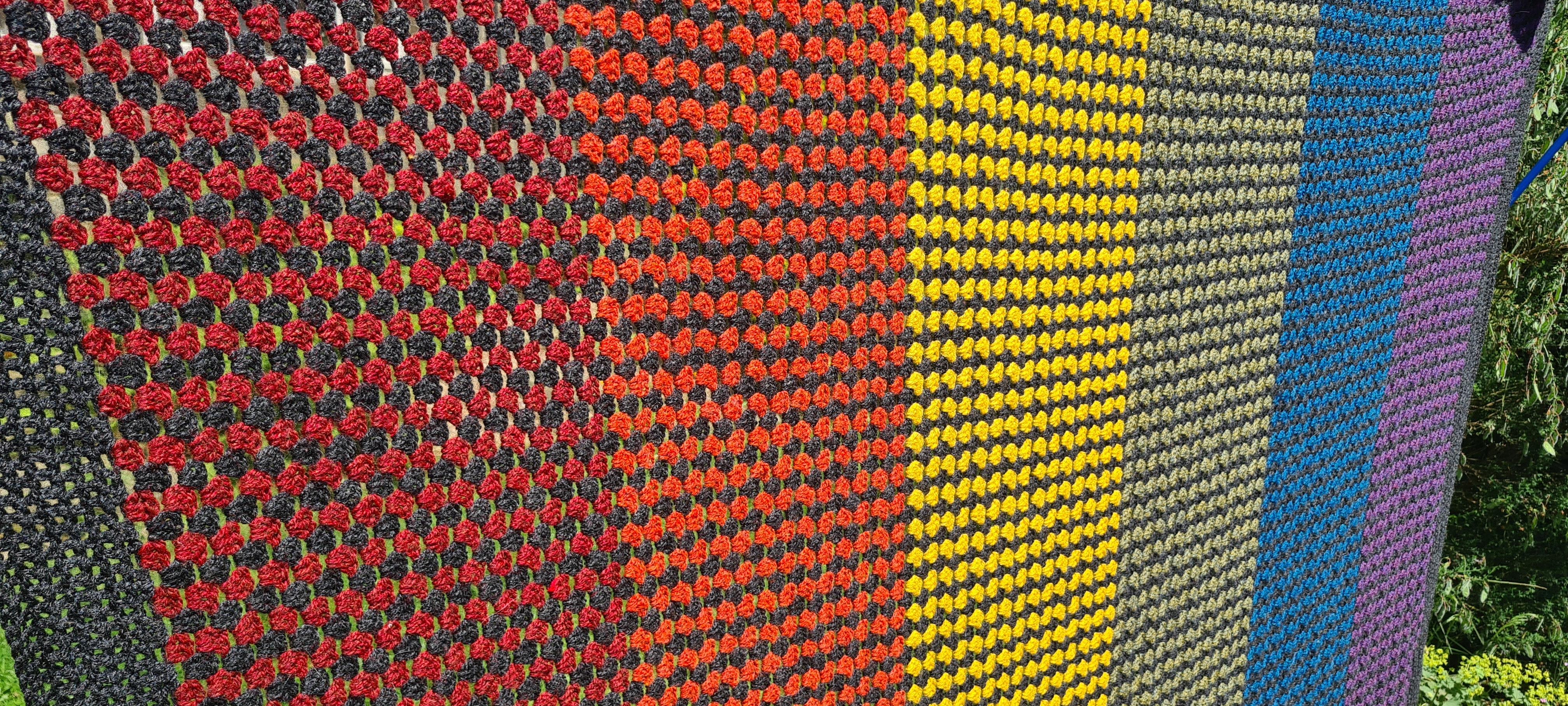 A relatively close-up shot of a crocheted blanket, made using the granny stripe technique. The blanket is a rainbow, made of six wide stripes (red, orange, yellow, sage green, blue and violet) but in-between each single row of colour, there is a row of a charcoal stitches. The border is charcoal too.