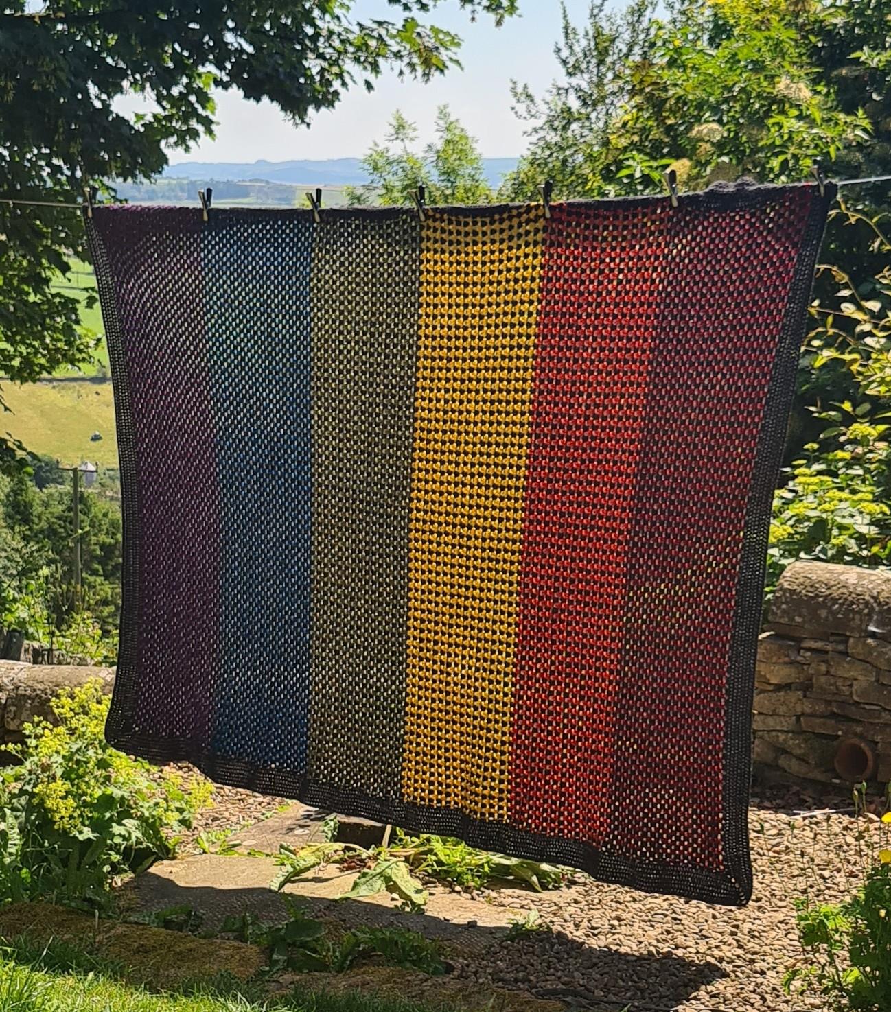 A wider shot showing the previously described blanket in full. It is hanging on a washing line in a garden, on a summer's day. The blanket is around 180cm by 240cm.