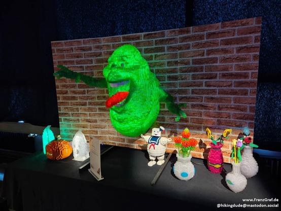This image showcases an impressive and creative LEGO display featuring a scene inspired by the Ghostbusters movie. The centerpiece of the display is a large, detailed LEGO model of the Stay Puft Marshmallow Man, emerging from a brick wall backdrop. The figure is vividly green with a red tongue, capturing the iconic character's playful yet menacing appearance.