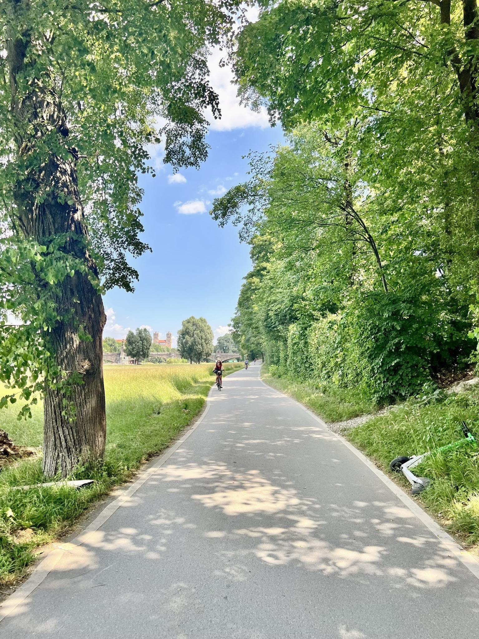 A paved path stretches forward, framed by tall, leafy green trees that cast dappled shadows on the asphalt. To the left, an open grassy field reveals a historic red-brick building with towers in the far distance under a bright blue sky. On the right, thick green hedges and trees line the path, with a green electric scooter resting on the grassy shoulder. A cyclist is visible riding in the distance.