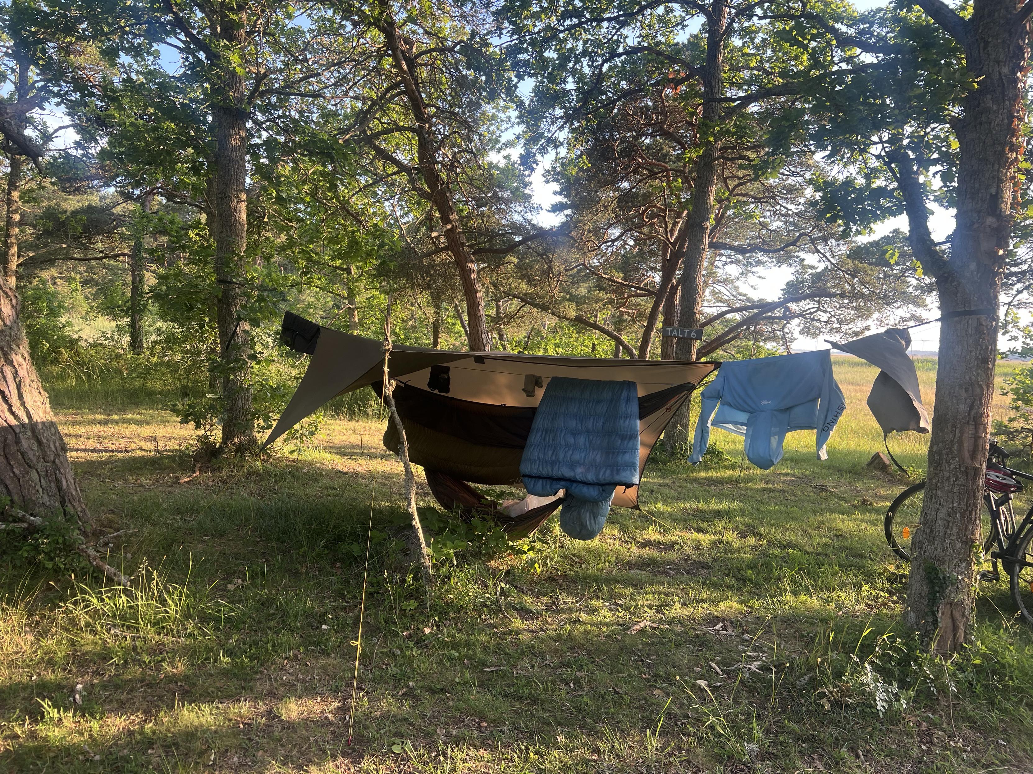 A typical hammock hang with a tarp pitched so one corner is lifted. Ridge line is used to air out clothes and quilt. There’s a gear hammock dangling underneath, too close to the ground.