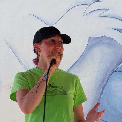 Woman with green old greenie fringe fest t-shirt wears black billed cap and speaks into microphone with blue and white art (not the dragon boat mural) behind here