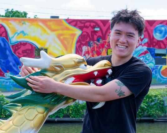 Man with dark shirt t-shirt and moderately short hair and a tattoo on his arm holds a golden and green dragon with a yellow red and blue mural in the far background