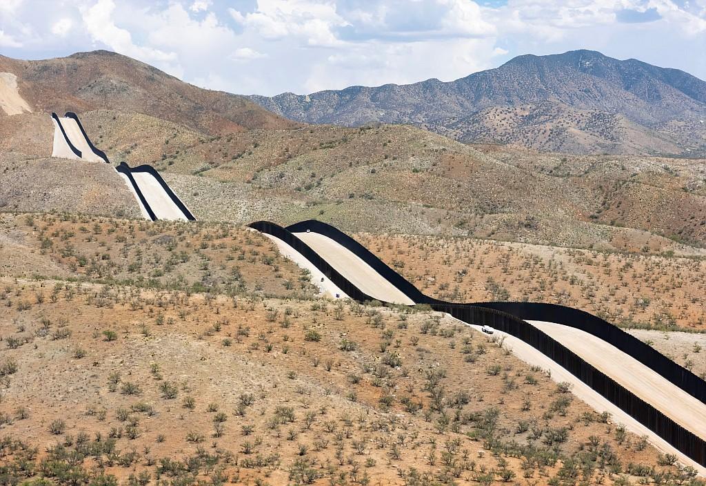 A wide, high-angle landscape shot showing a tall, dark metal border fence snaking dramatically across an arid, hilly desert terrain in Arizona. The fence is constructed of tightly spaced vertical steel bollards that cast dark shadows on a wide, unpaved dirt patrol road running directly alongside it.

The undulating barrier cuts over multiple ridges and valleys, undulating across the dry hillsides dotted with sparse, low-lying desert shrubs and cacti. In the lower right half, a white vehicle is visible on the patrol road. The background opens up to rolling desert hills and distant rugged mountains under a blue sky filled with scattered, bright white clouds.