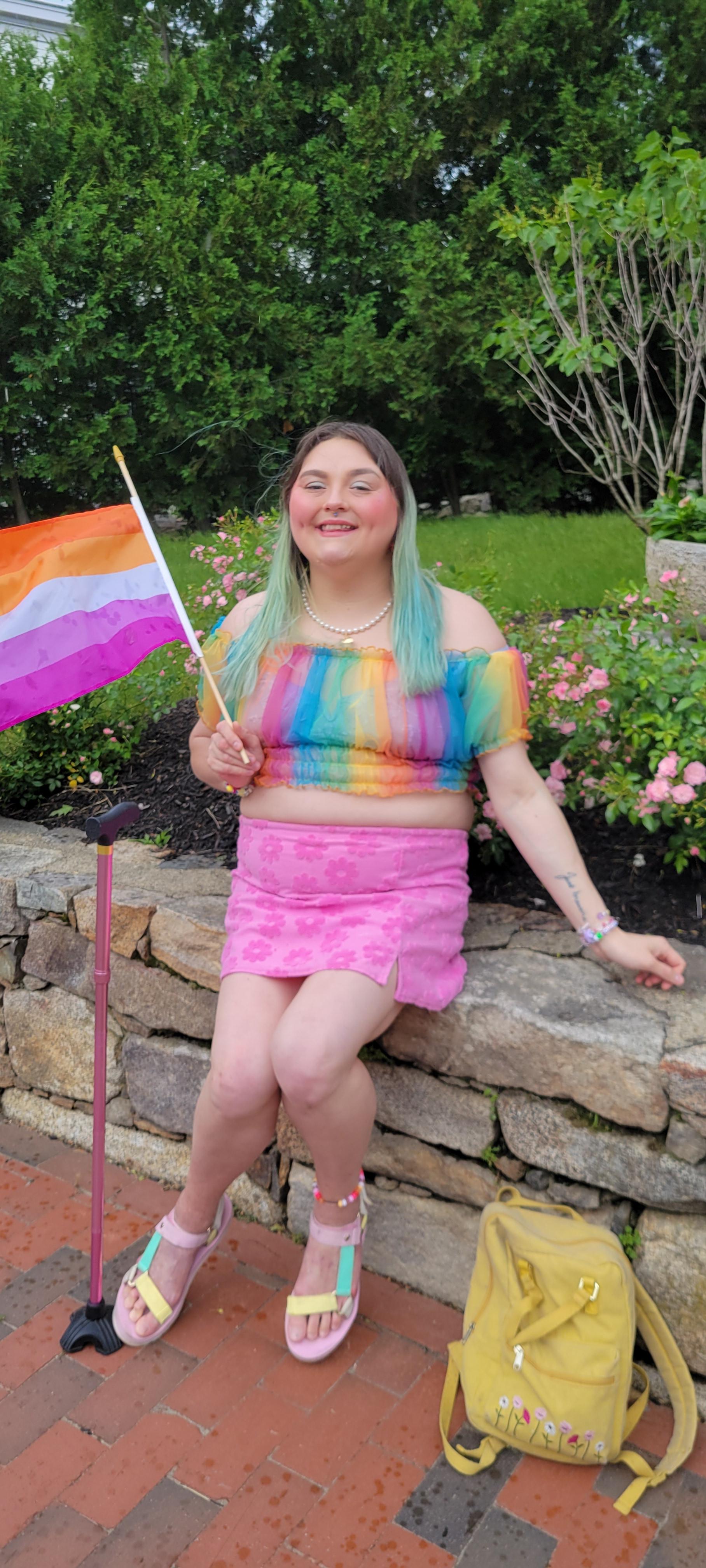Me sitting on a rock wall I am wearing a pink skirt with a rainbow mesh shirt, Holding the lesbian pride flag.. My pink cane is standing next to me and I am smiling