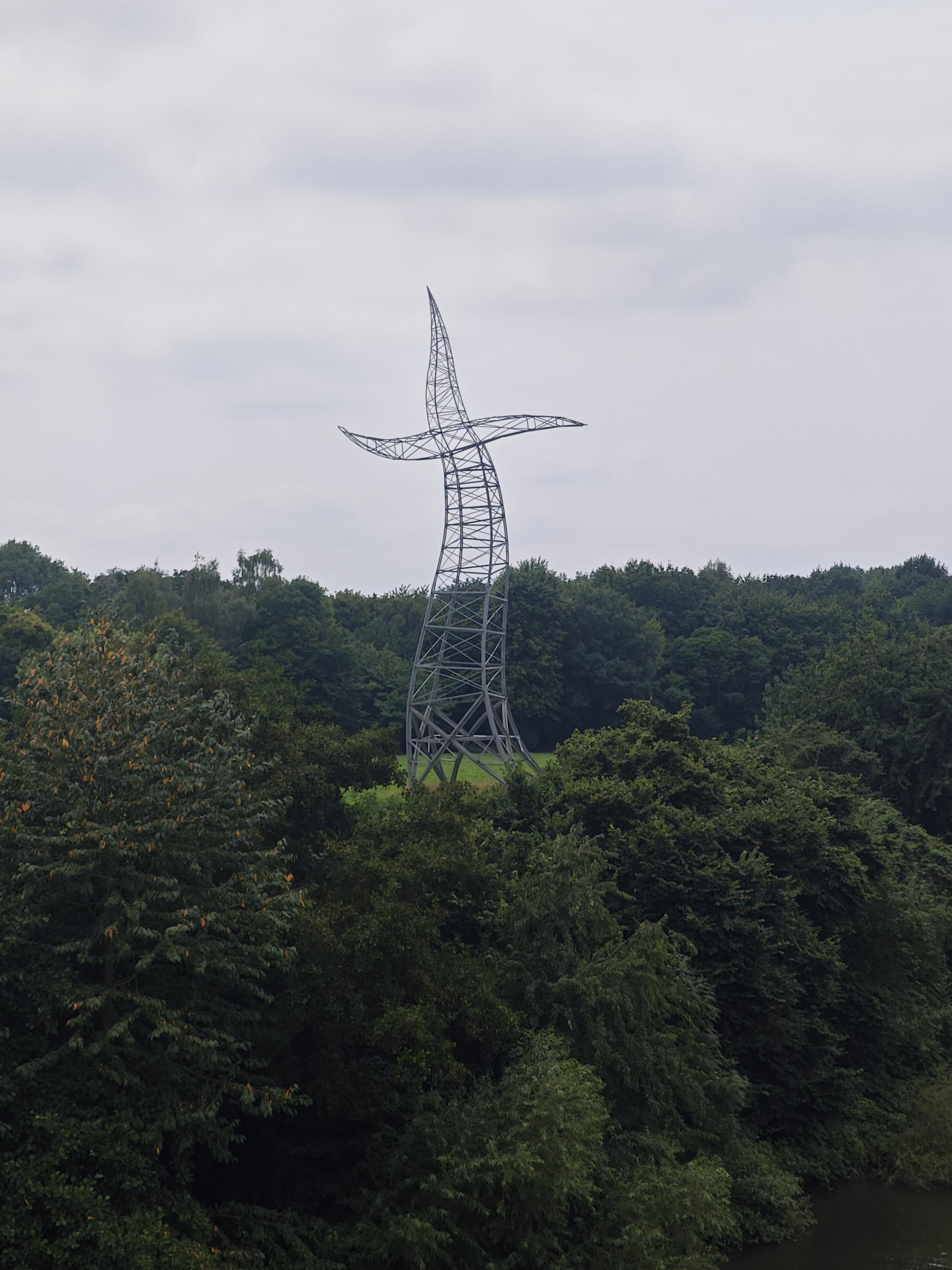 Bild einer Skulptur, die so ähnlich wie ein Strommast aussieht.
Durch die geschwungene Form sieht die Skulptur aus, als wäre sie in Bewegung.
Rund herum sind grüne Bäume zu sehen.
Der Himmel ist leicht grau und bewölkt.
