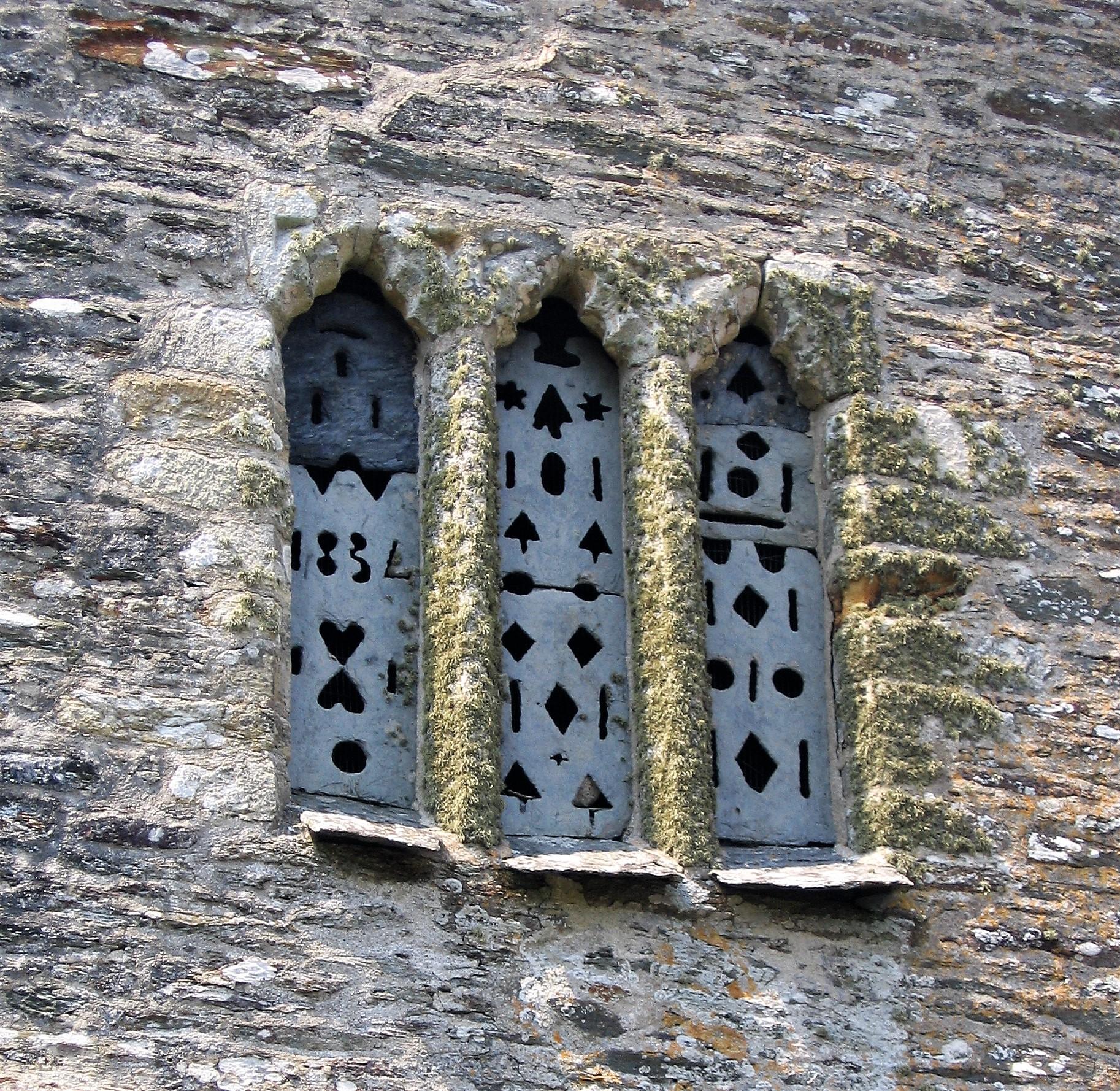 A window in a church wall filled with carved pieces of slate to provide ventilation. There are three sections divide by stone mullions. The carvings are a mix of hearts (upright and inverted), circles, diamonds, lines, stars and tree-like shapes. In the left hand section the number 1834 is carved to denote the date. The wall around the window is made of slate-type stone, while the mullions and window surround look like they may be granite, most of which is covered in lichen. The tops of the mullions are finely carved.