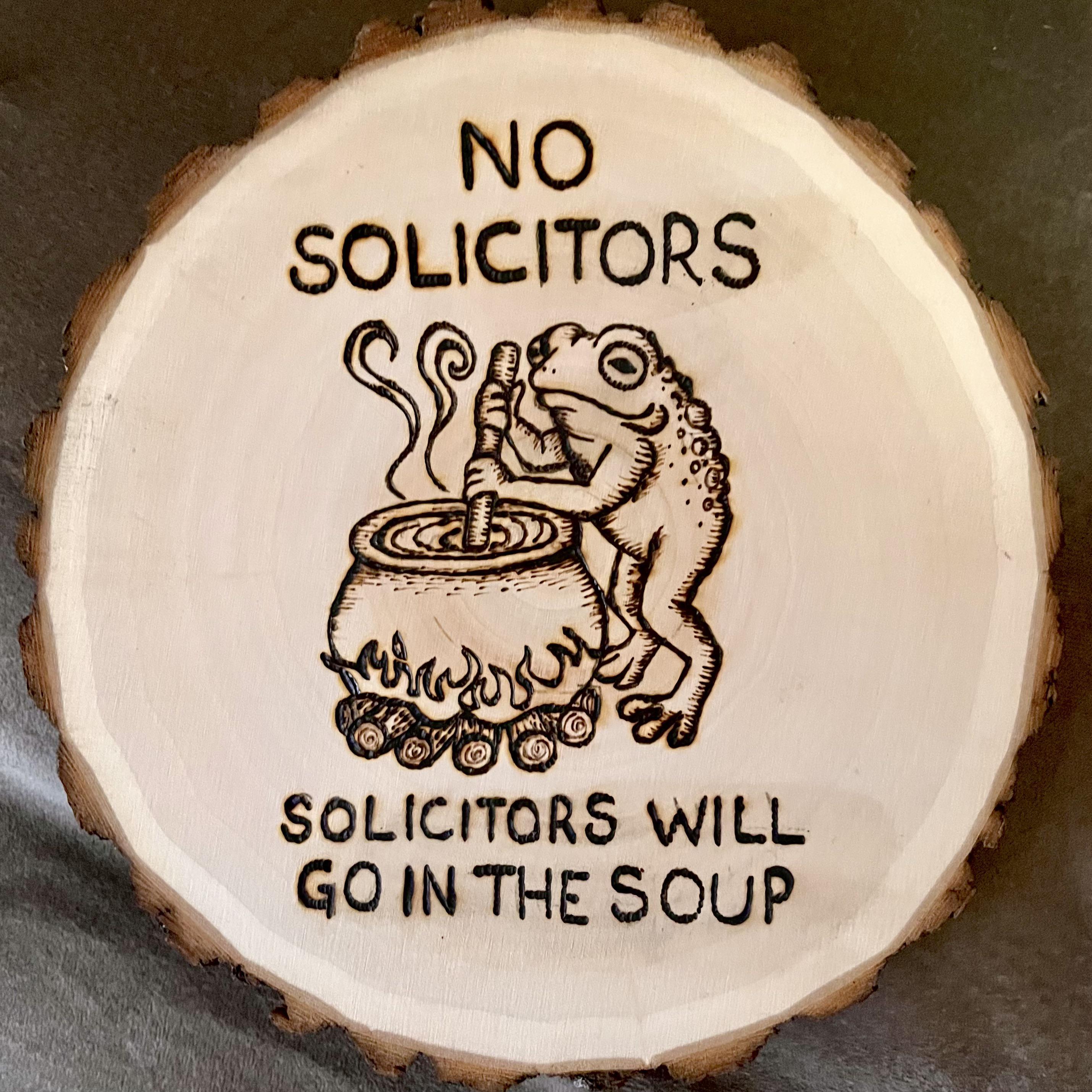 Wood burn art on a log cross section. A frog stands upright, stirring at a witch's cauldron atop a fire. Caption: No solicitors. Solicitors go in the soup.