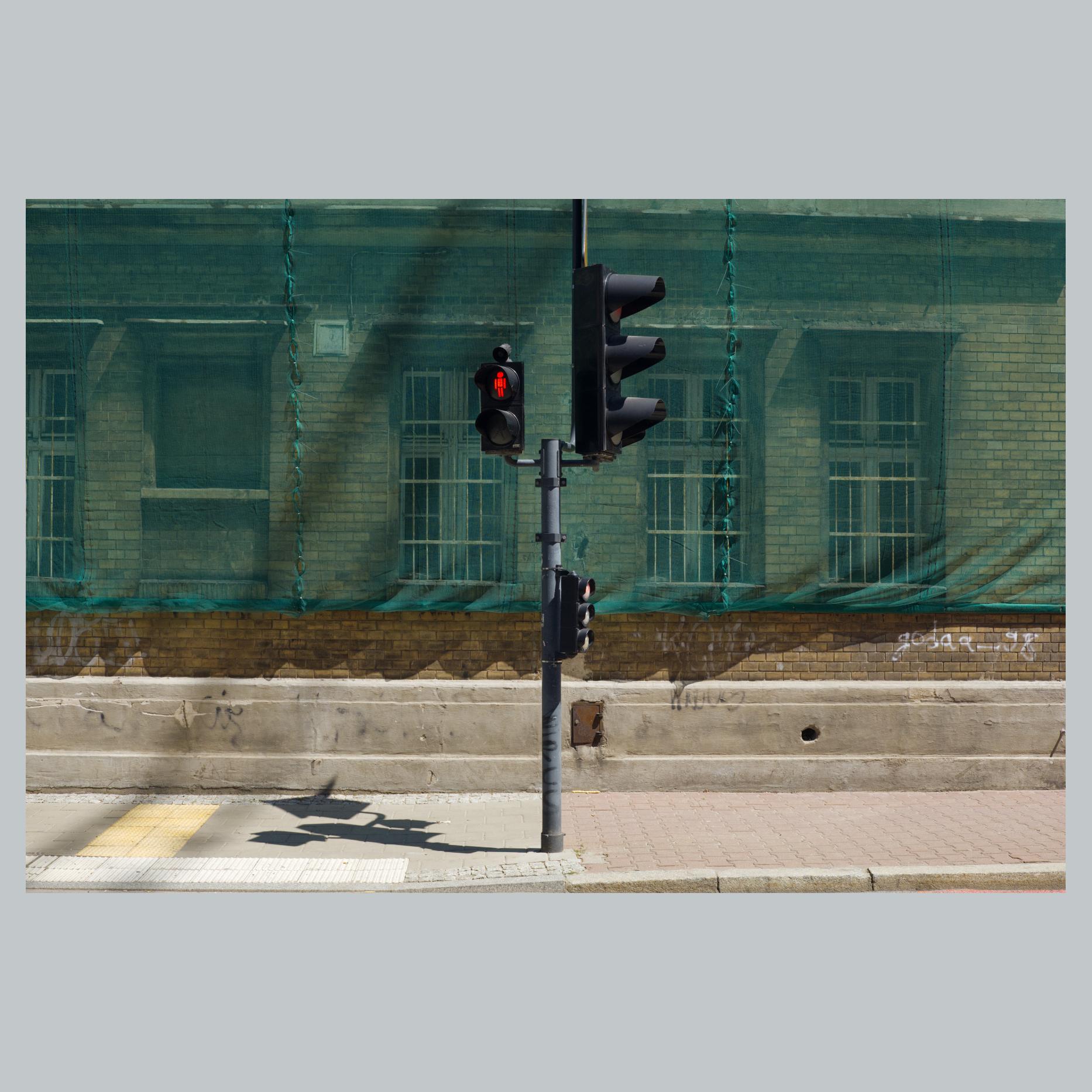 Na fotografii widać stojący w centrum kadru sygnalizator świetlny, który ze wszystkich stron wyświetla czerwone światło. Stoi na oświetlonym ostrym światłem chodniku. Tło dla niego stanowi budynek z zieloną płachtą. Jest środek dnia.