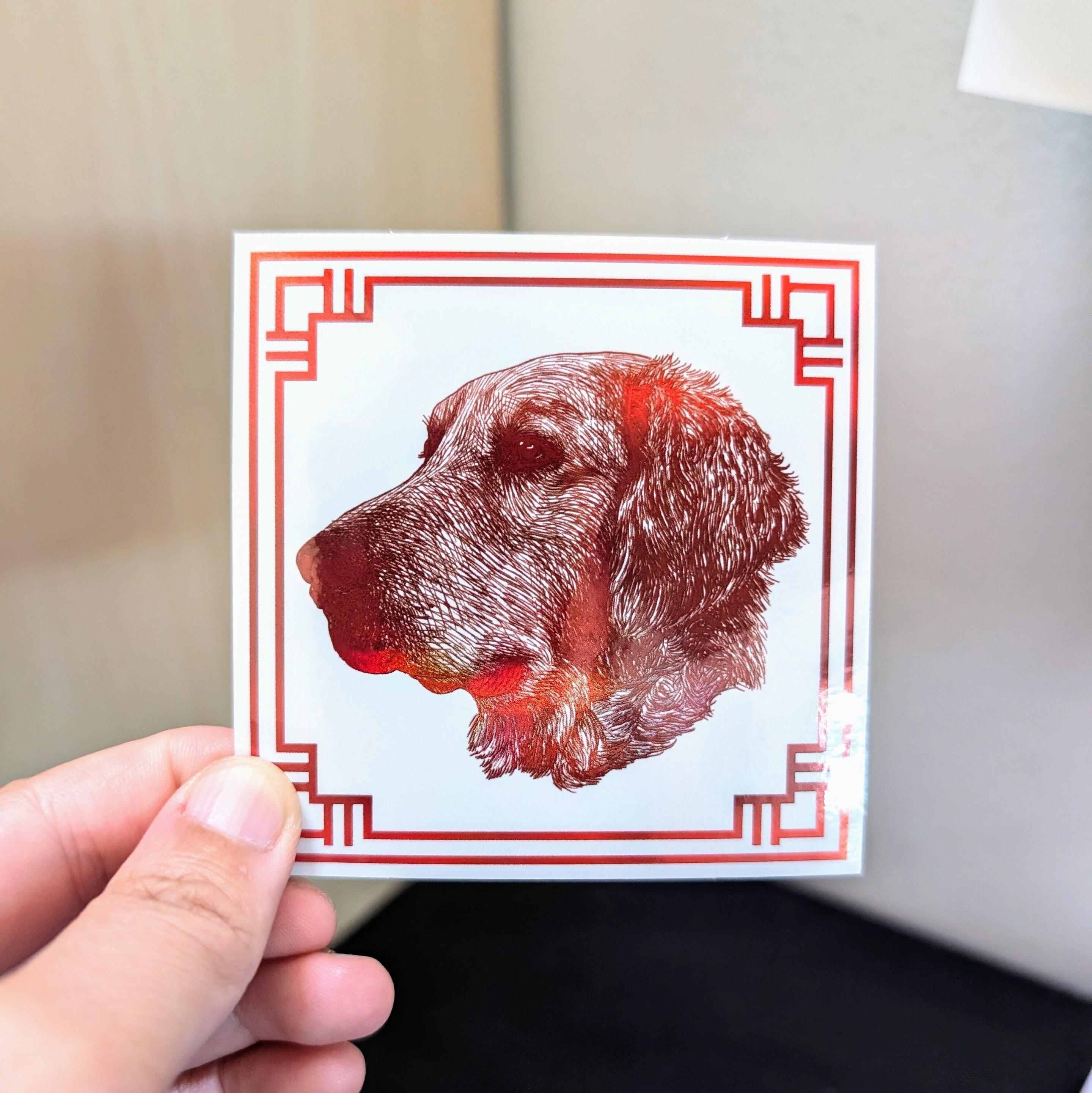 Photo of a left hand holding a square sticker. The sticker is a holographic drawing of the Chinese Zodiac animal: the Dog.