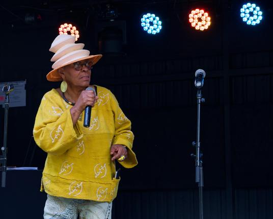Black woman wears yellow shirt and cylindrically symmetrical pleated hat with stage lights behind