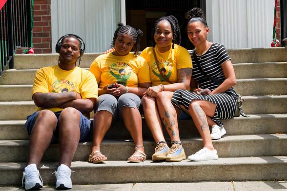 Four black people sit on the steps,  a black man wearing headphones with his arm crosseded, two woman with braided hair and a woman wearing a horizontal black and white striped dress,  the others all wear yellow t-shirts.