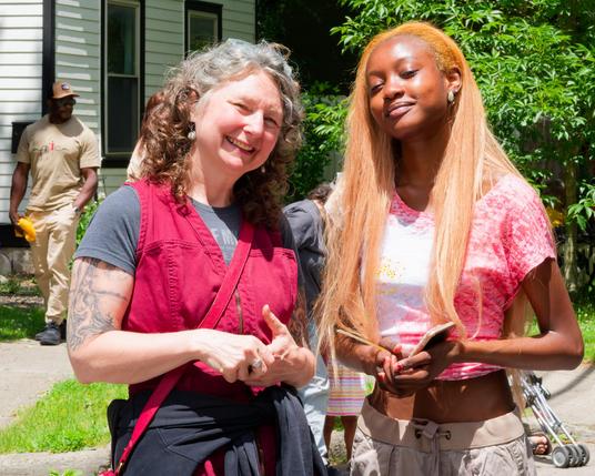 On the left a white woman with mostly grey hair and to the left a black lady with blonde hair with a house and a big bush or tree in background