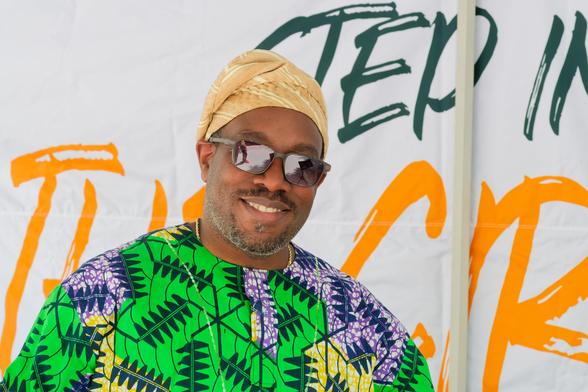 Black man wears scarf on head and geometric yellow black green and white shirt and wears shades with a sign behind him that reads (partially visible) step into the circle.