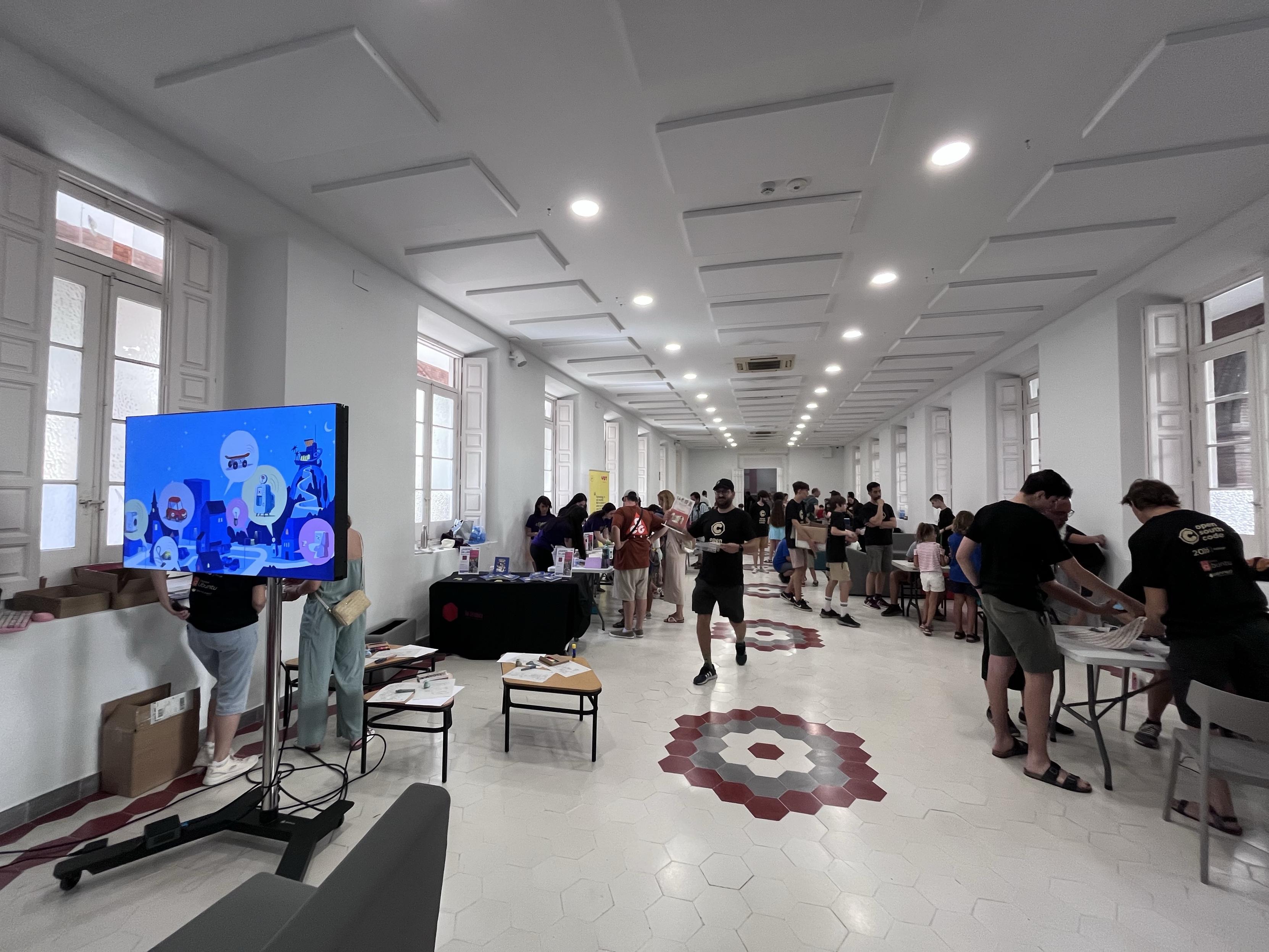 An interior space with a large group of people engaged in various activities, including tables with items on display and a TV screen showing animated graphics.