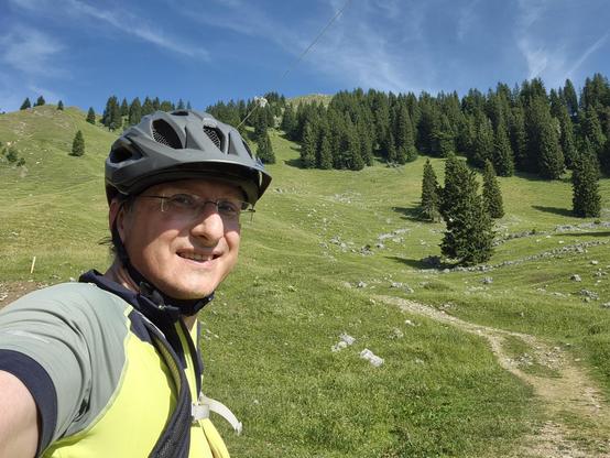 This image captures a joyful and scenic moment during a cycling or mountain biking adventure. In the foreground, a person is taking a selfie, wearing a black helmet, glasses, and a bright yellow and gray cycling jersey. The individual is smiling, reflecting the enjoyment and excitement of the ride.
The background reveals a stunning mountainous landscape with rolling green hills dotted with evergreen trees. A winding dirt trail cuts through the meadow, leading the eye deeper into the scene. The …