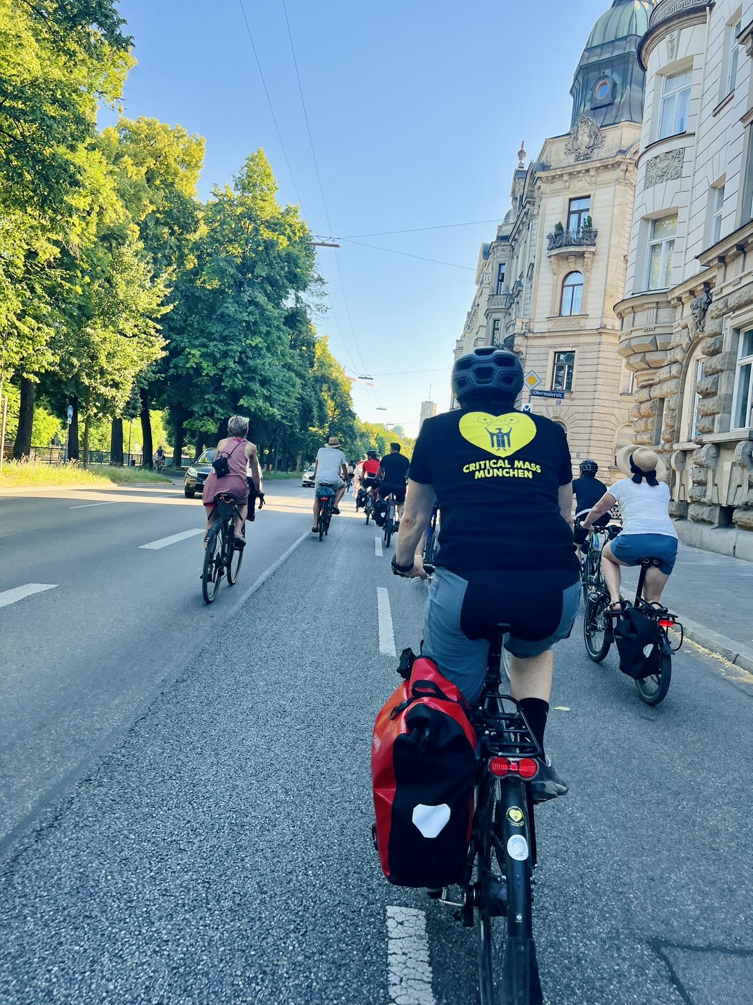 A group of cyclists rides along a wide city street on a sunny day. In the foreground, a cyclist wearing a black helmet and a black t-shirt with a yellow "Critical Mass München" heart logo rides a bicycle with a red pannier bag. Lush green trees line the left side of the road, while elegant historic apartment buildings border the right under a clear sky.
