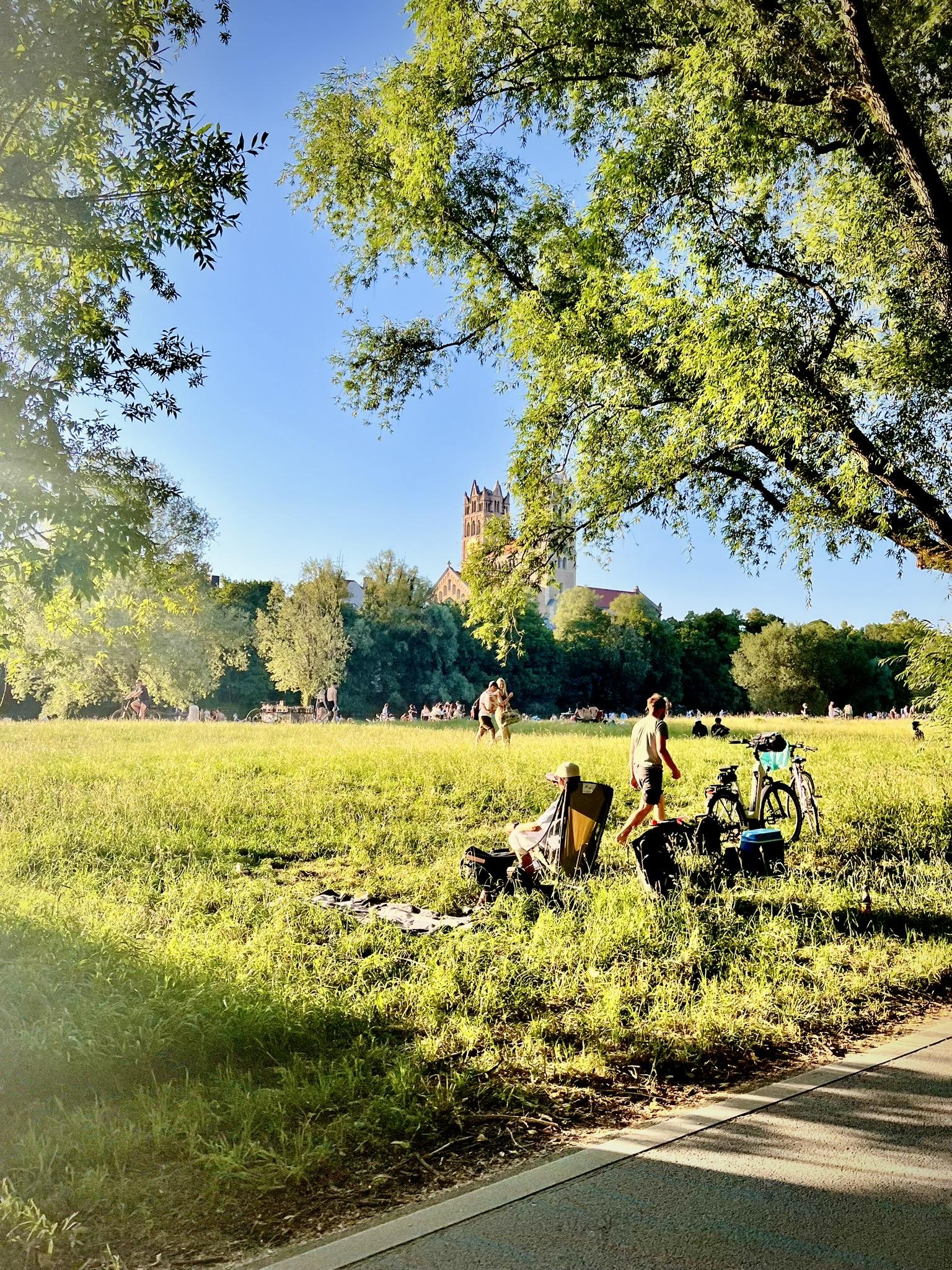 A sunlit grassy park under a clear blue sky, framed by lush green tree branches in the foreground. In the meadow, people relax and walk, including one person seated in a folding chair next to parked bicycles. In the background, a historic church with brick towers rises above a line of dense trees.