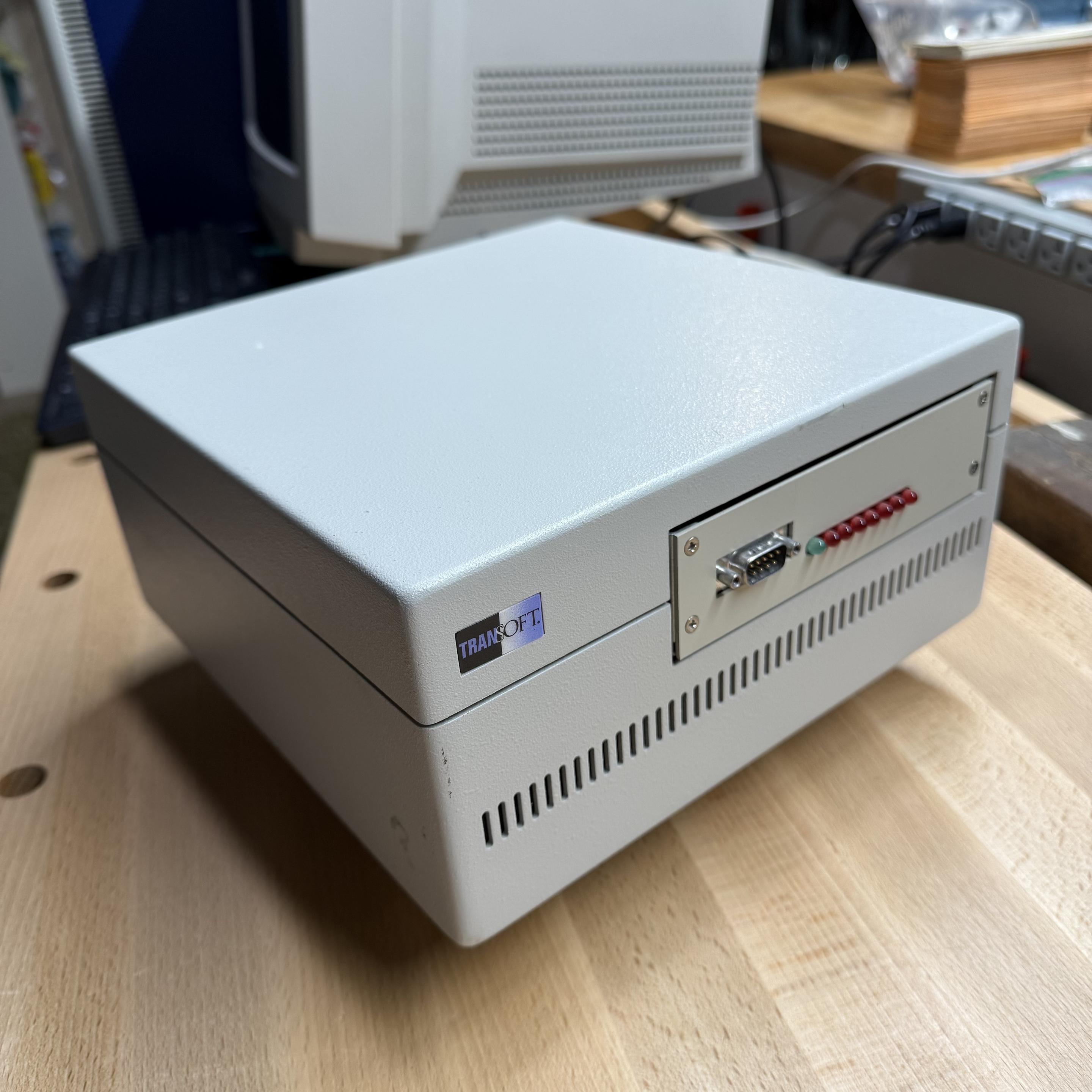 A piece of computer equipment in a grey metal case sitting in a wooden workbench. The front of the case has a sticker reading “TRANSOFT”, a DE-9 connector, a row of LED indicators, and a series of vertical ventilation perforations.