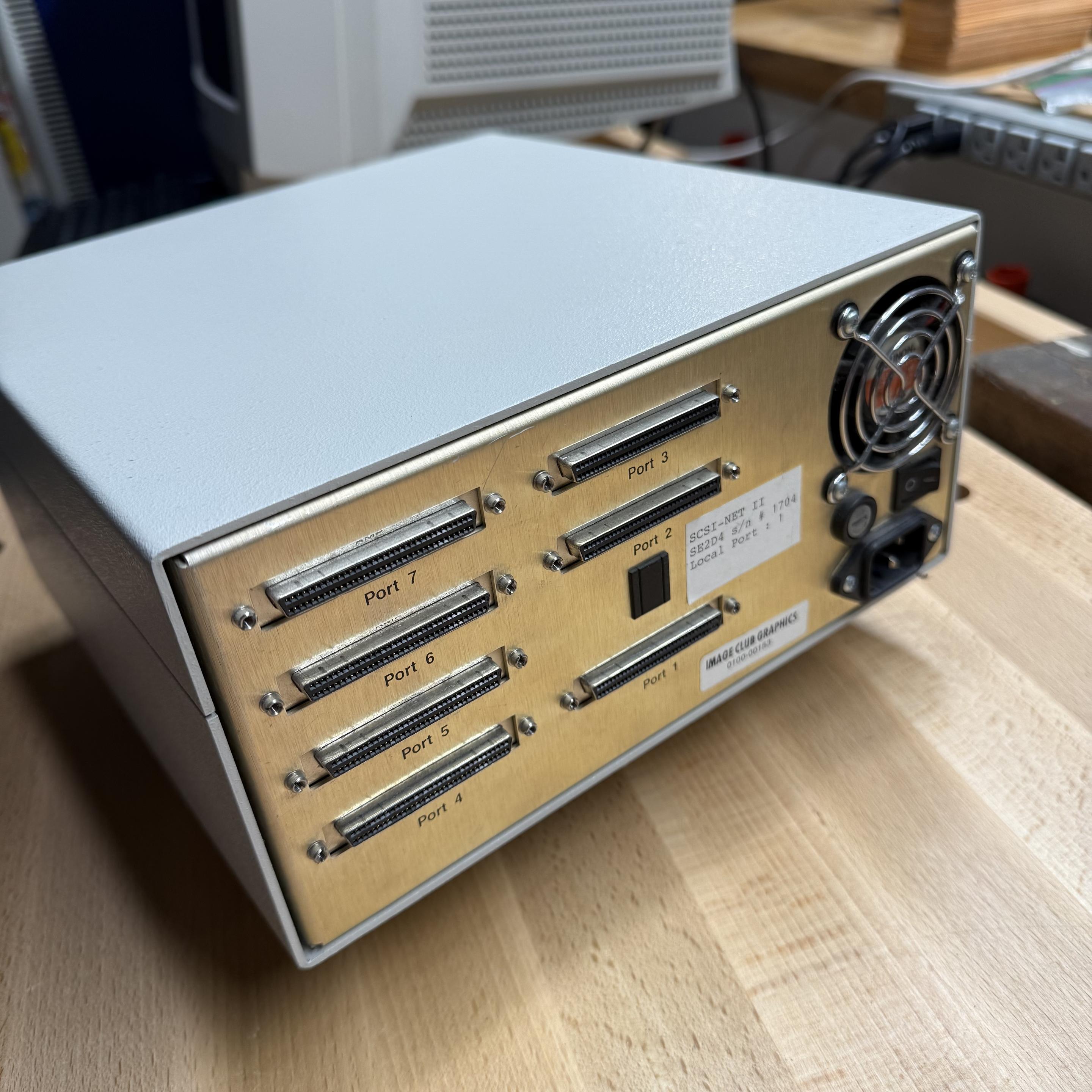 A piece of computer equipment in a grey metal case sitting in a wooden workbench. The back of the case has a pair of stickers, a fan grill, seven SCSI connectors, a power switch, and a power connector.

One sticker reads:

SCSI-NET II
SE2D4 s/n 1704
Local Port : 1

A second sticker reads:

IMAGE CLUB GRAPHICS
0100-00183