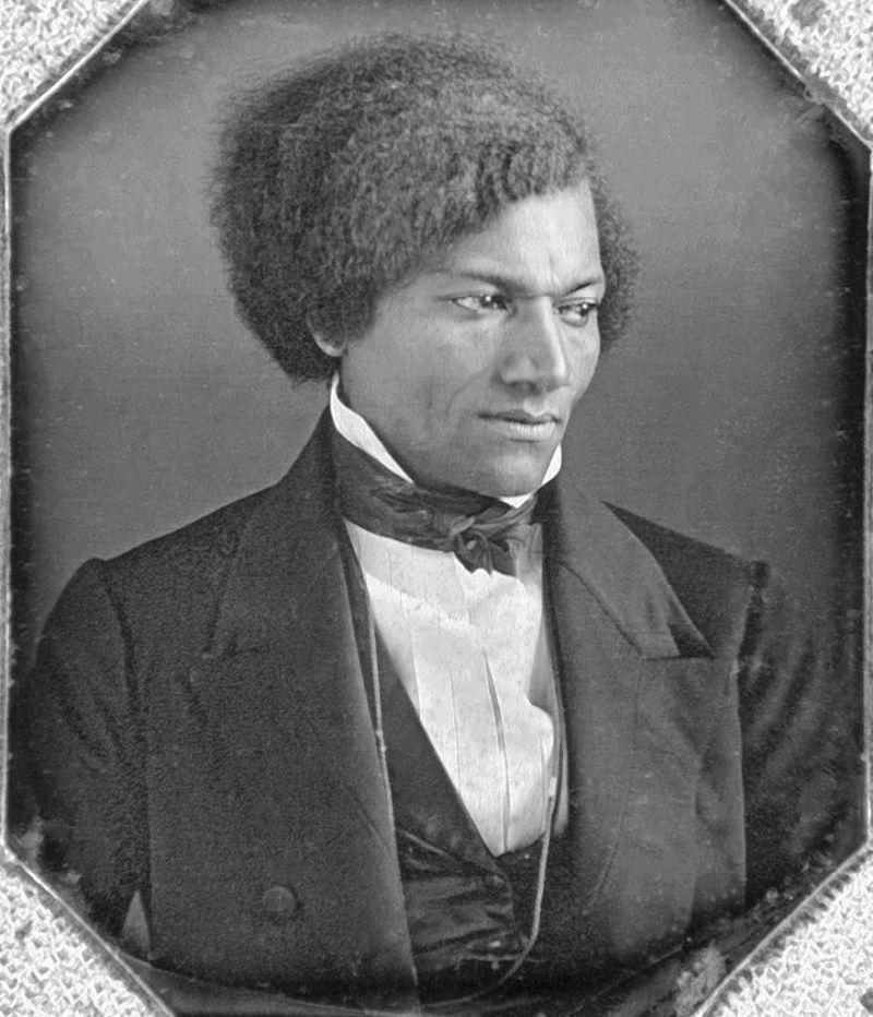 Photograph of a young Frederick Douglass (1840). He is looking thoughtfully down to his left. His hair is full, dark, and voluminous afro-textured. It is parted on his left.  He is wearing a dark formal suit jacket with a matching dark waistcoat.  Around his high-collared white dress shirt he has a  large, dark cravat tied in a bow.