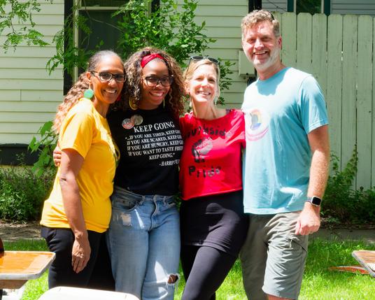 Three woman (left) and one man (right), pose together in front of sidewalk, house and treelawn