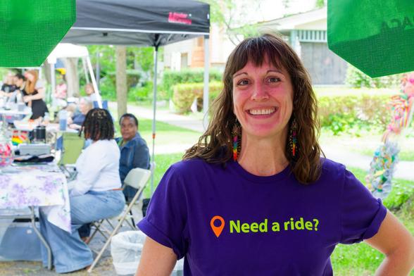 Woman wears purple shirt with a location icon and the text "Need a ride?" with shoulder length hair and a big smile (she's looking at a fox!) and more people can be seen behind tabling under tents