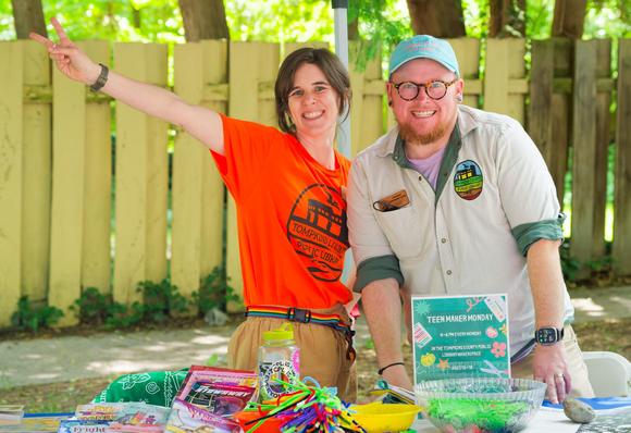 Woman in orange shirt with rainbow belt and beige pants holds up arm diagonally and makes v-sign as man in beige short-sleeved shirt, blue cap and glasses leans in,  in front of them is assorted stuff on a table and behind them a yellow fence with irregular boards,  maybe the kind that tom sawyer got his friends to whitewash