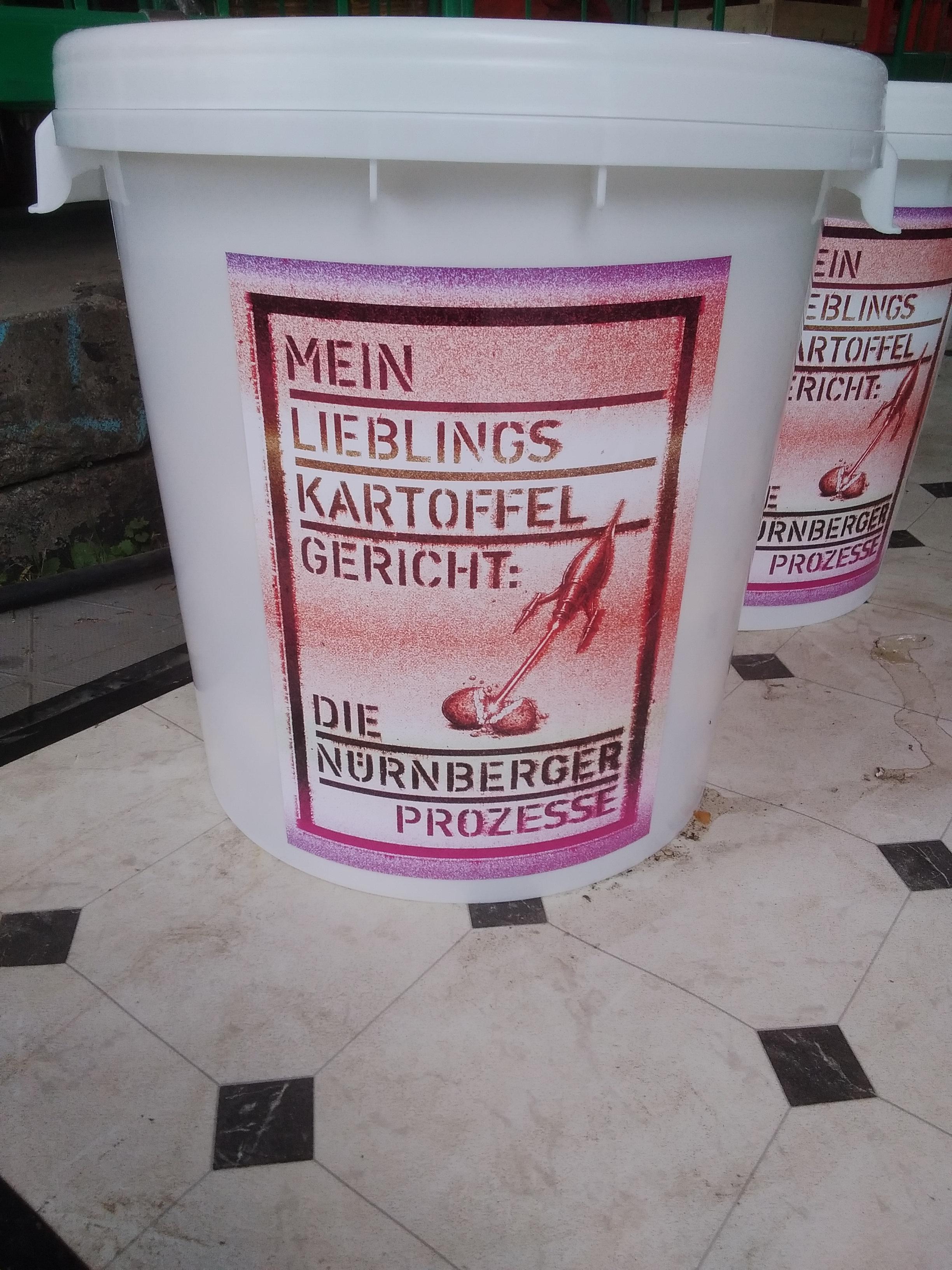 Big white buckets for foods standing on a bit dirty white and black linoleum floor, a message printed on them: "Mein Lieblingskartoffelgericht: die Nürnberger Prozesse" ("My favourite potato dish is the Nuremberg Trials").