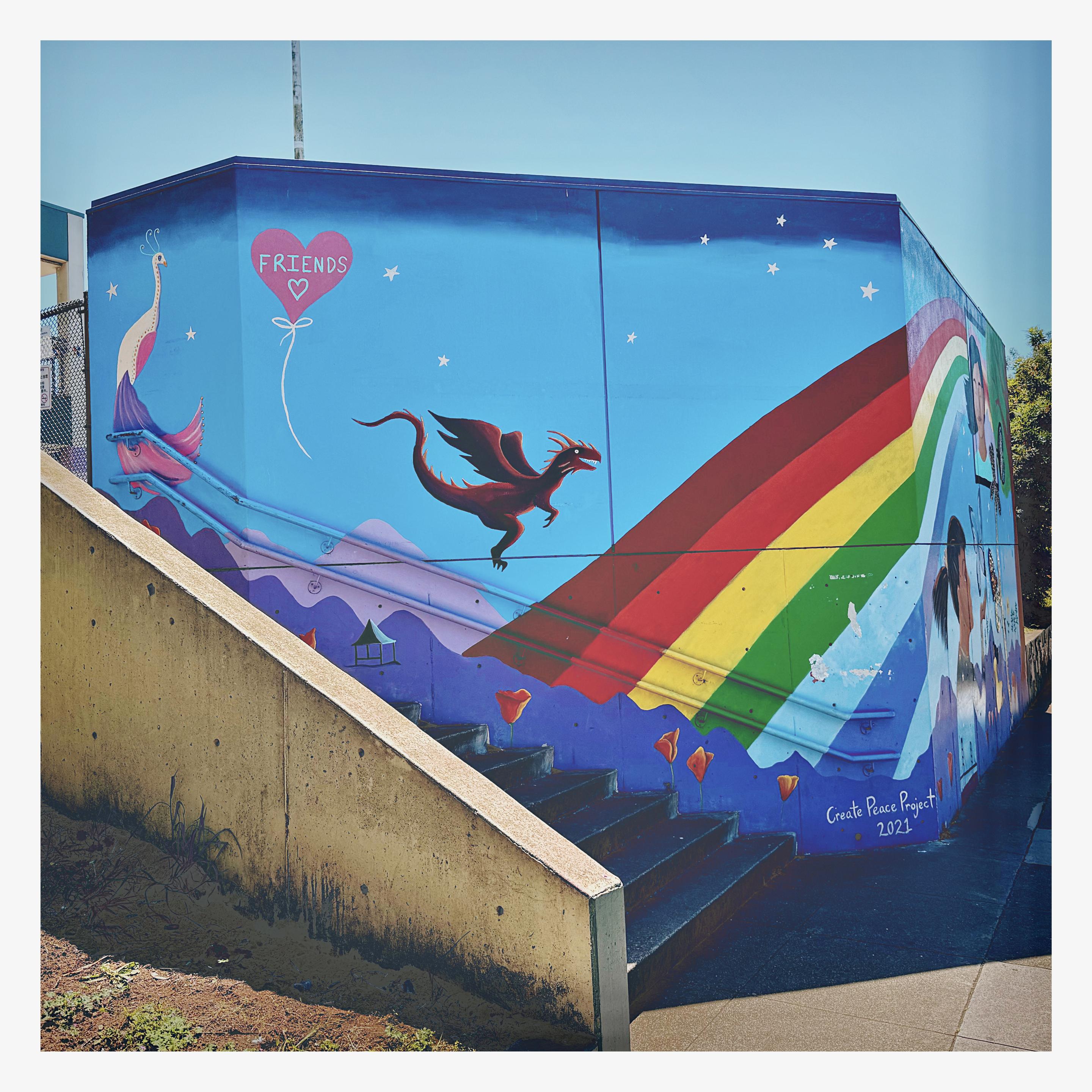 Photo of a joyful mural painted on a large concrete wall. We are looking at small smiling red dragon flying over purple mountains with a gigantic rainbow in the background. There is a small pink balloon floating above the light blue skies that reads “friends 🤍” next to a beautiful white, pink, and purple peacock. Along the bottom are painted orange California poppies and as the mural continues around the side of the wall there are images of smiling kids and adults along with some butterflies and trees. In front of the mural is a concrete staircase leading up to Monroe Elementary School. Taken in San Francisco, in the Excelsior.