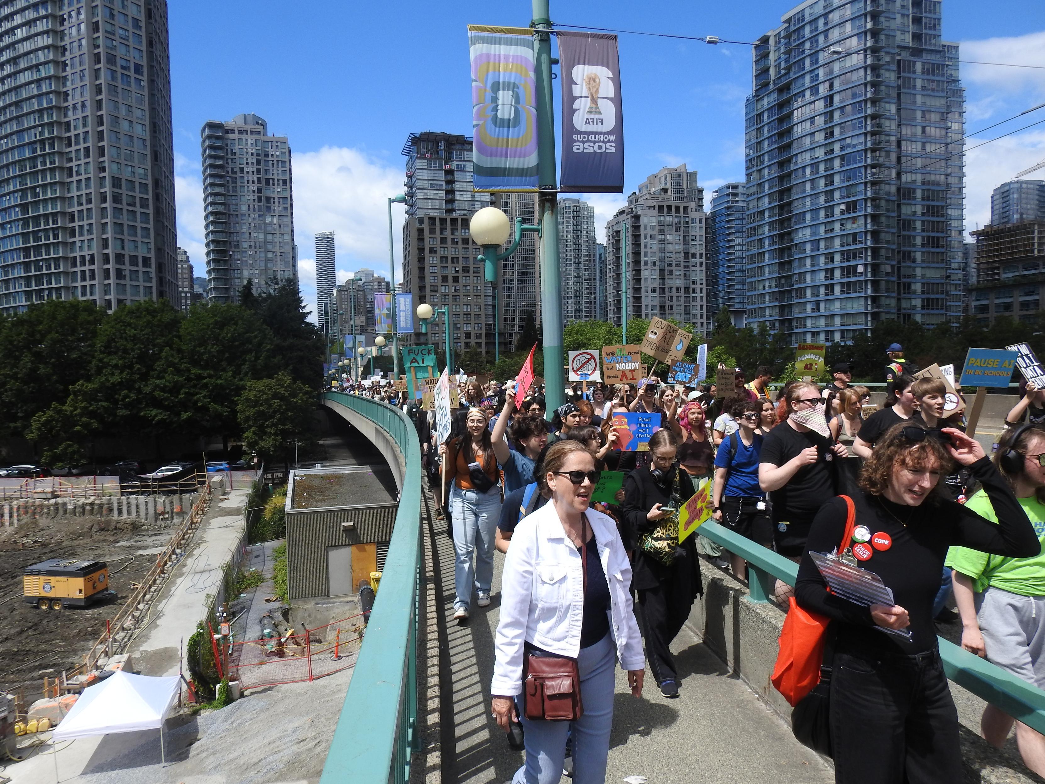 A photo of a long column of around a thousand protesters crossing Cambie Bridge in Vancouver, with several skyscrapers in the background. The crowd stretches beyond the visible part of the bridge. Many of the protesters are carrying signs critical of AI.