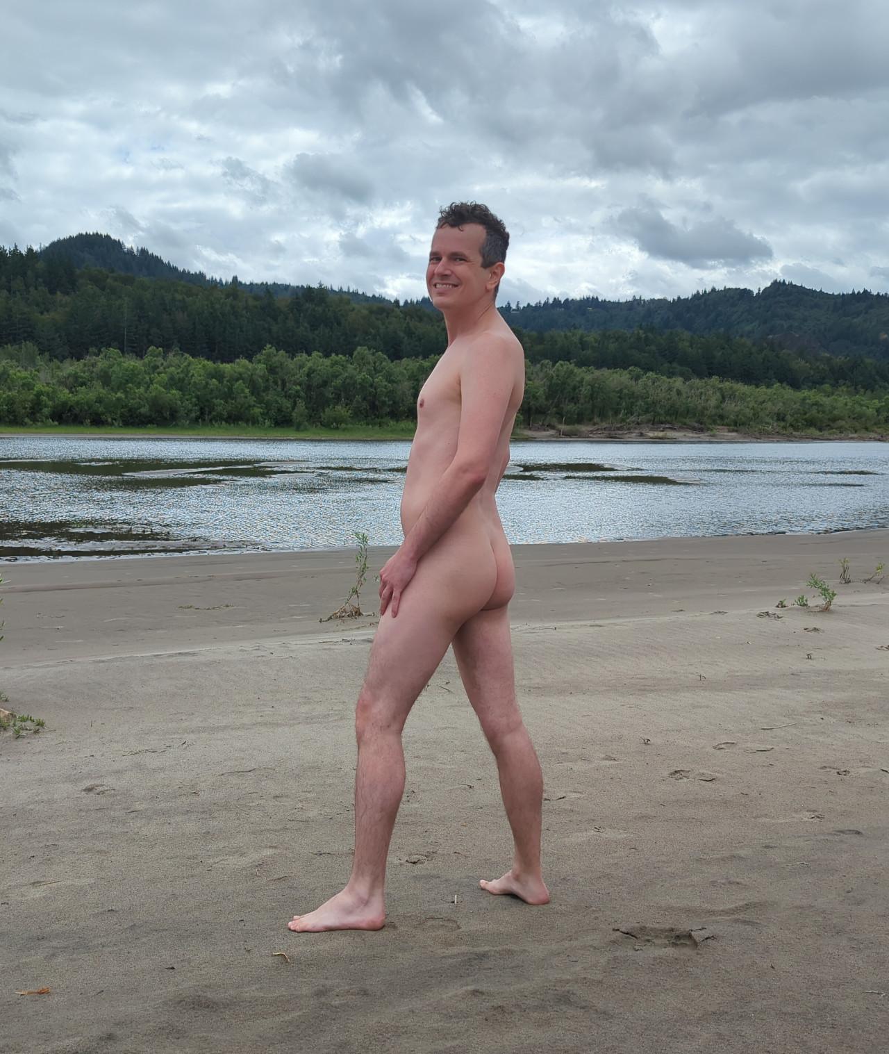 A nude photo of me standing barefoot on the sandy shore of the nude beach at Rooster Rock State Park. I am standing in nearly side profile toward the left of frame, arms relaxed at my sides with my backside and bare bubble butt visible, while my face looks at the camera with a grin. Hills covered in trees line the opposite shore across the Columbia River with a grey cloudy sky.