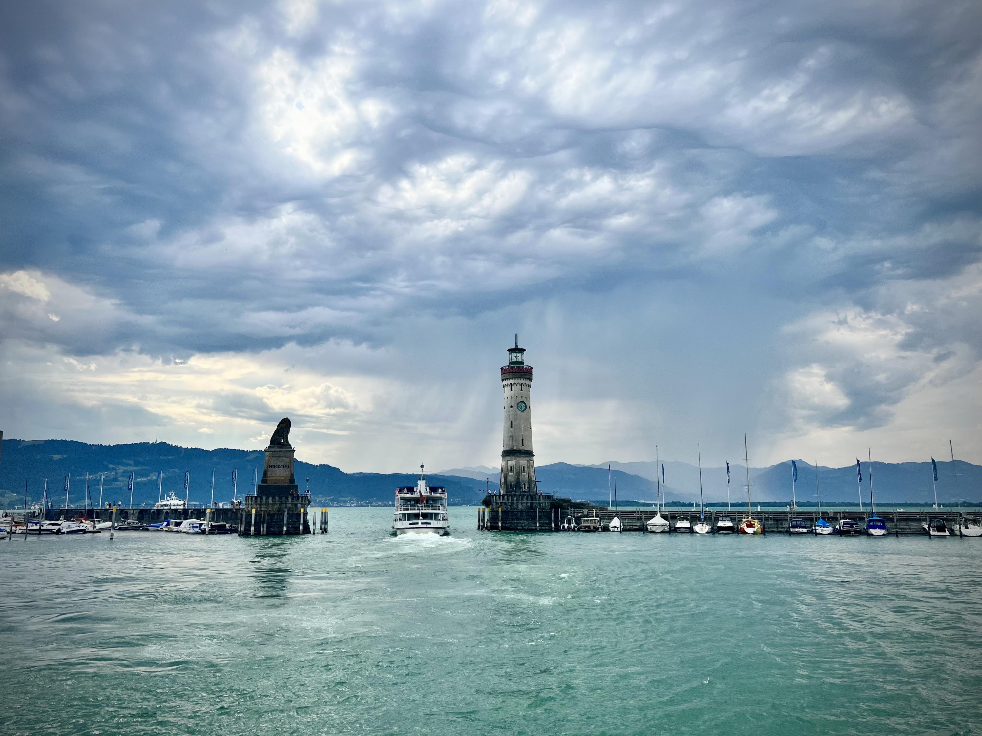 A white ferry boat sails through the entrance of a harbor, framed by a historic stone lighthouse on the right and a large stone lion statue on the left. Docked sailboats line the piers on both sides of the entrance, set against the backdrop of a vast turquoise body of water, distant mountains, and a dramatic, cloudy sky.