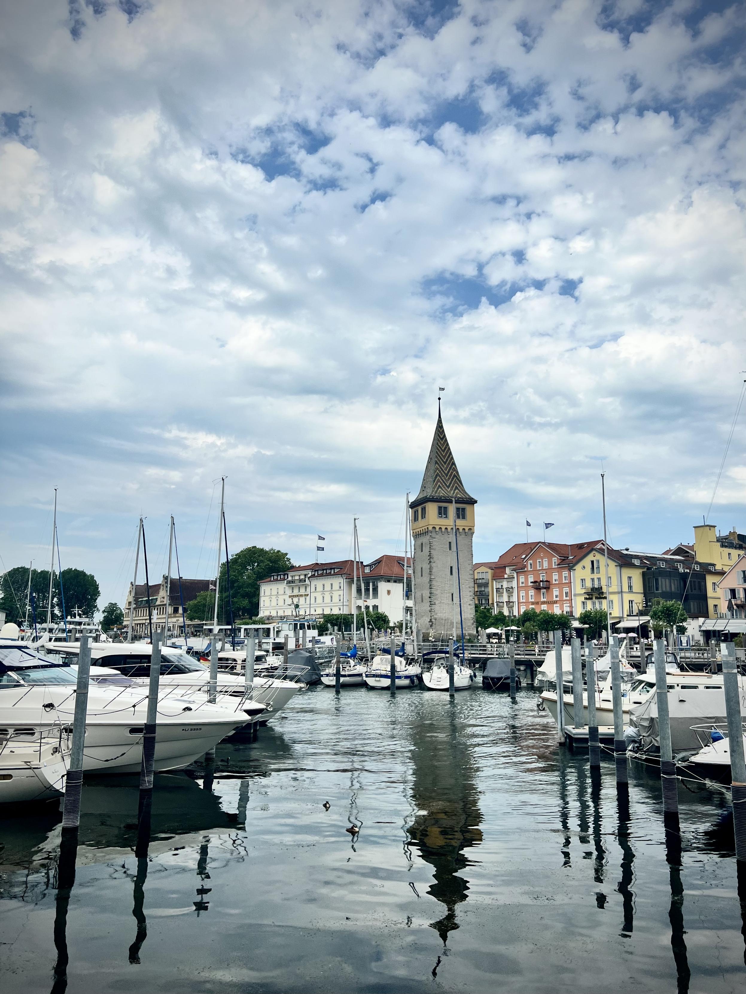 A medieval stone tower with a steeply pointed, colorful tiled roof stands prominently on a waterfront, reflected in the calm harbor water below. Numerous white yachts and sailboats are docked in the foreground, while a row of pastel-colored, multi-story buildings lines the shore under a sky filled with dramatic clouds.