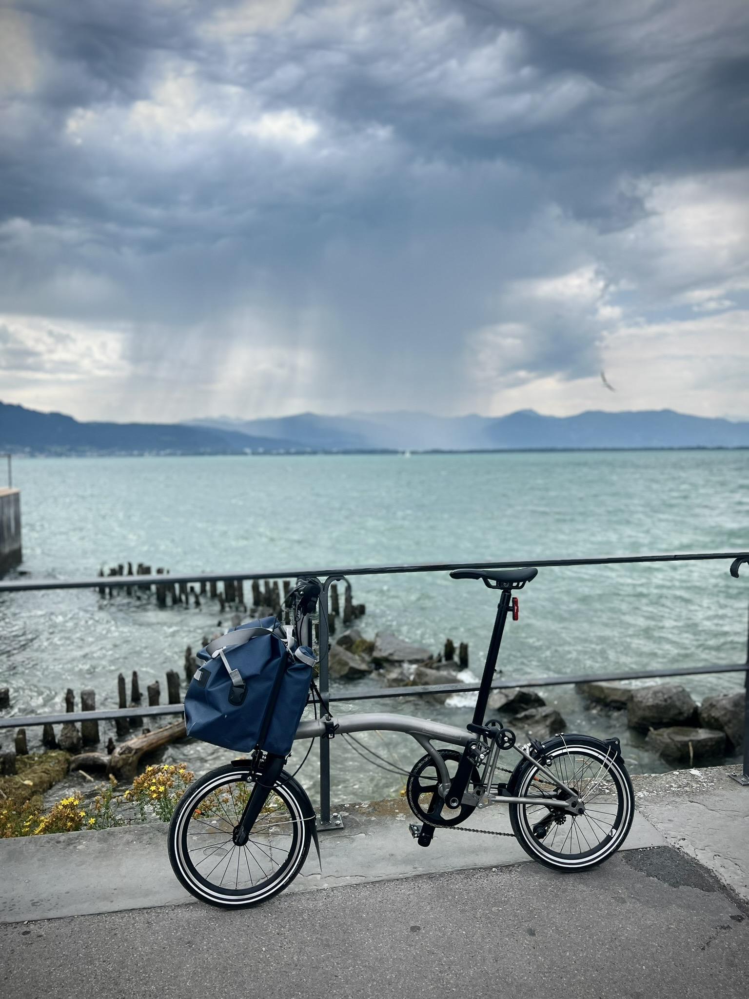 My titanium-grey Brompton with a large blue bag mounted on the front frame is parked on a concrete path next to a black metal railing. Behind the railing, rocky shores and wooden pilings meet a vast expanse of turquoise water, while in the far distance, sunbeams break through dark storm clouds over a mountain range.