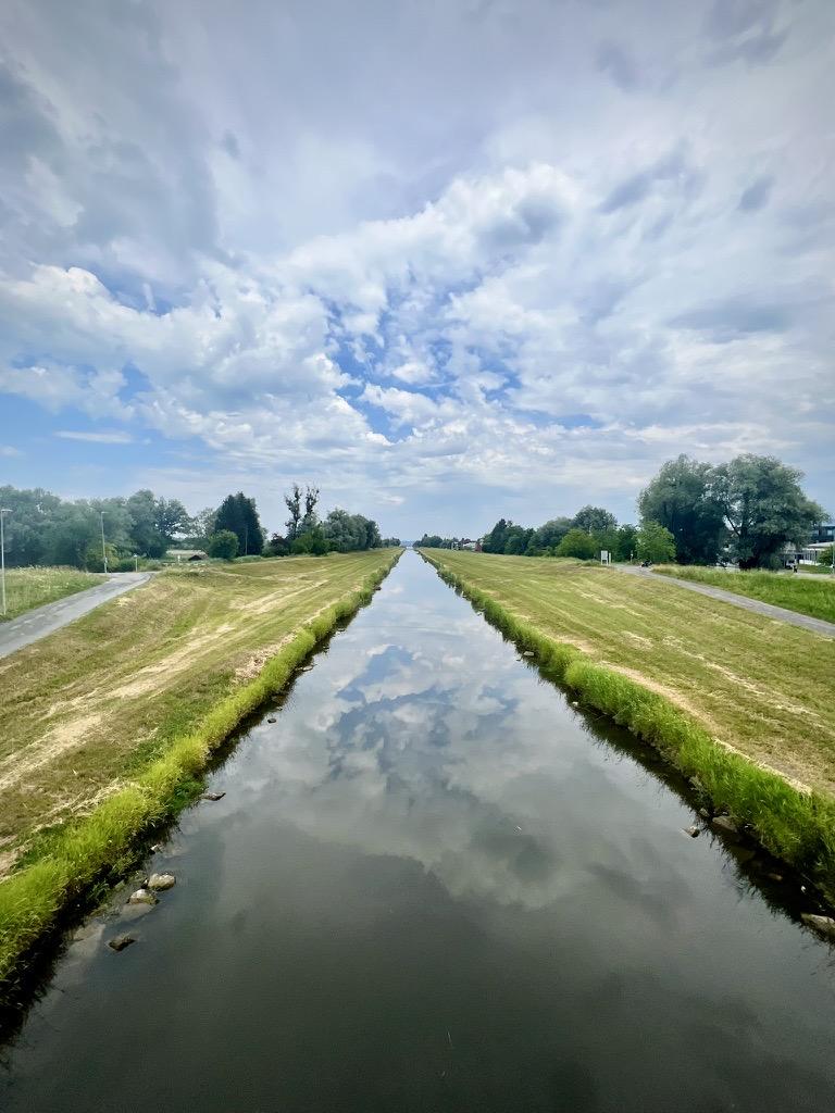 A perfectly straight canal stretches symmetrically toward the horizon, flanked by grassy banks and paths on either side. The calm, mirror-like surface of the water reflects a vibrant blue sky filled with dramatic, white and grey cloud formations.