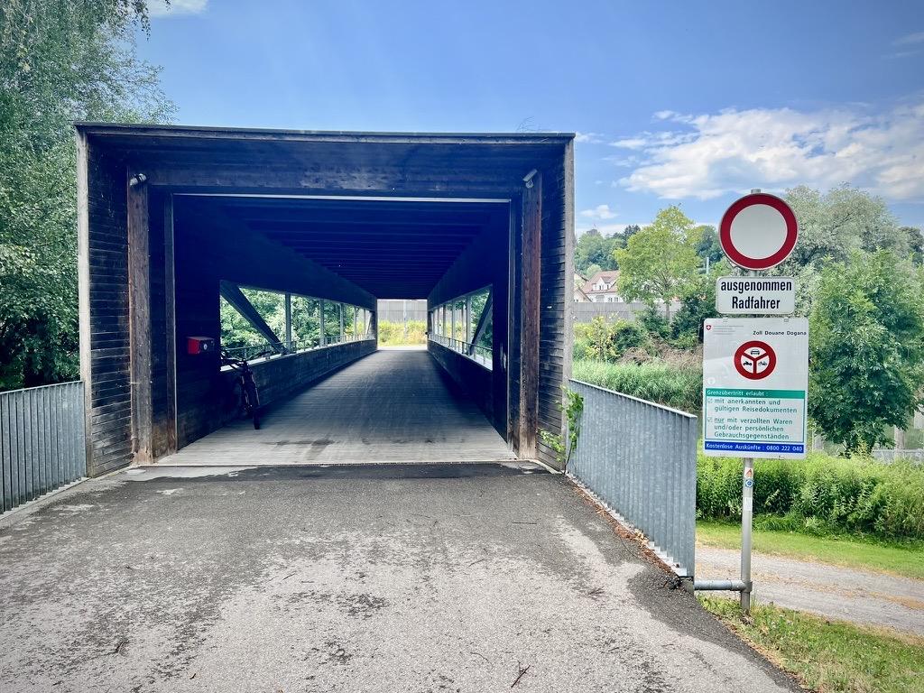 ‎⁨Gaißau⁩, ⁨Vorarlberg⁩, ⁨Austria⁩ 

The entrance of a covered wooden bridge with dark horizontal siding opens onto a paved path. To the right, a metal signpost displays a red-bordered "no vehicles" traffic sign with the German text "ausgenommen Radfahrer" below it, alongside a larger white customs informational sign, while a bicycle rests parked inside the bridge's shadowed wooden interior.