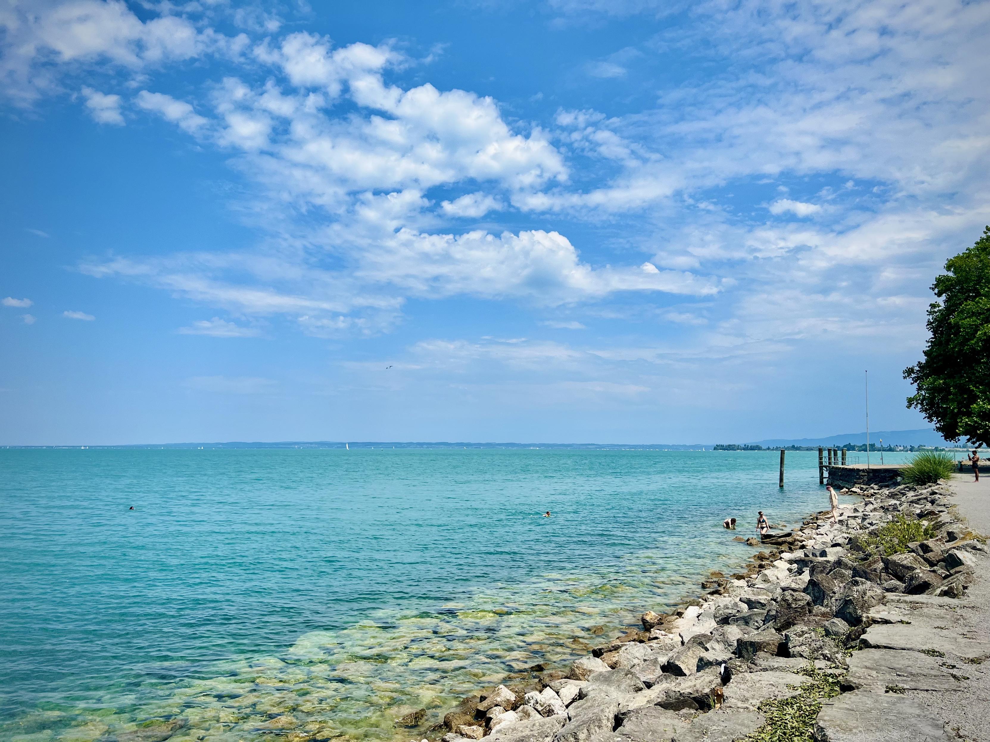 A bright lakeside scene with a wide expanse of calm, turquoise-blue water stretching to the horizon. In the foreground right, a rocky shore forms a natural edge, with smooth stones and a narrow path.

Small groups of people sit and stand along the rocks near the waterline. A pair of wooden posts protrude from the water, and a low dock or platform extends from the right side.

Above, the sky is clear and vivid blue, Lake Constance, filled with large, fluffy white clouds. In the distance, faint shoreline silhouettes and gentle hills are visible, adding depth to the landscape.