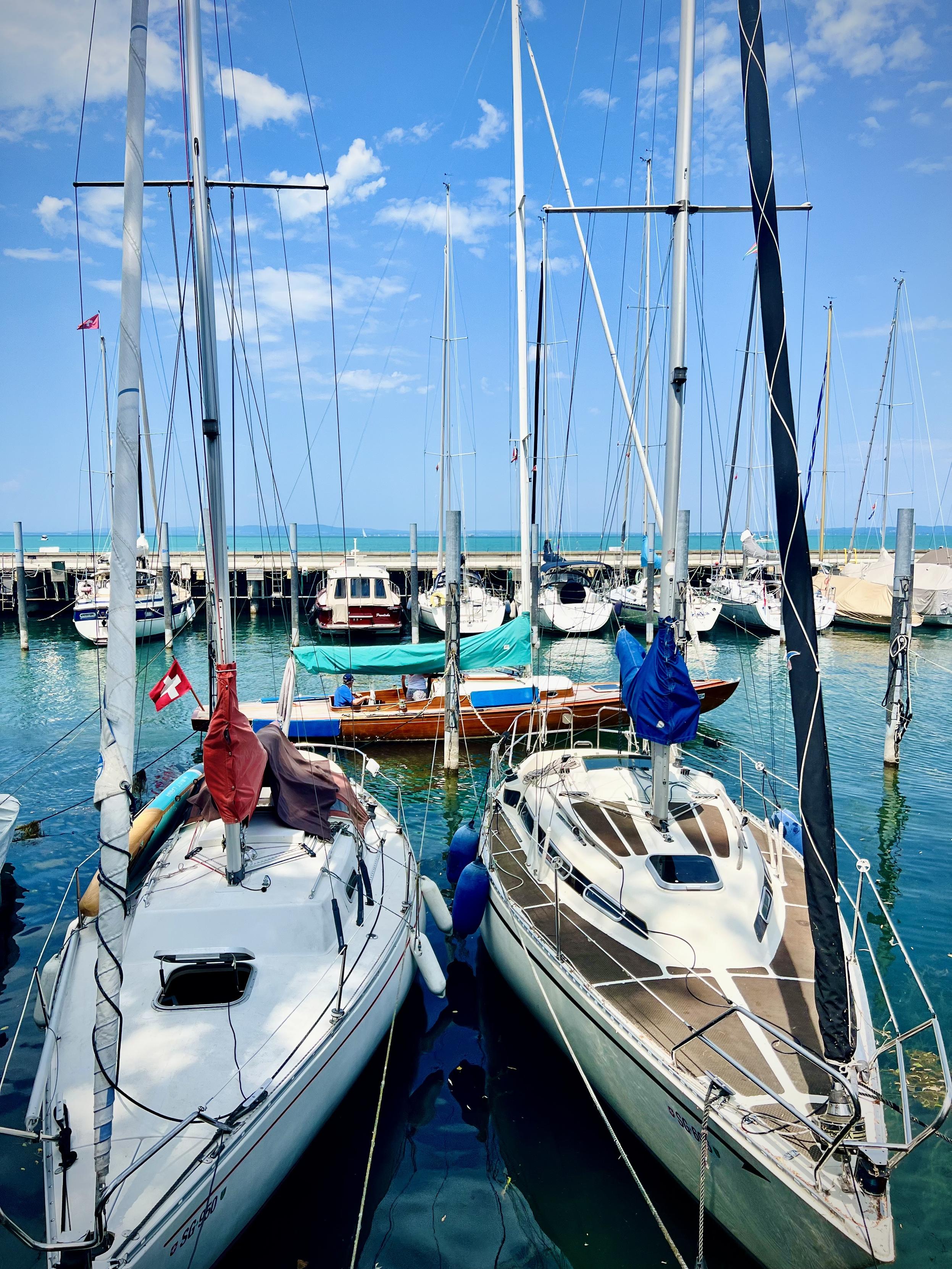 A tranquil marina scene on a sunny day, filled with sailboats moored at wooden docks. In the foreground, two white yachts sit side-by-side; the left one features a small Swiss flag and a red sail cover, while the right one has a sleek, dark mast and brown deck detailing.

Behind them, several other boats—ranging from smaller wooden vessels with turquoise covers to larger yachts—are gently floating in the clear, blue-green water. Vertical wooden pilings rise from the harbor to keep the boats secured. In the distance, a long concrete pier stretches across the frame, separating the marina from the expansive, turquoise lake beyond. The sky is a vibrant blue, dotted with light, wispy white clouds.