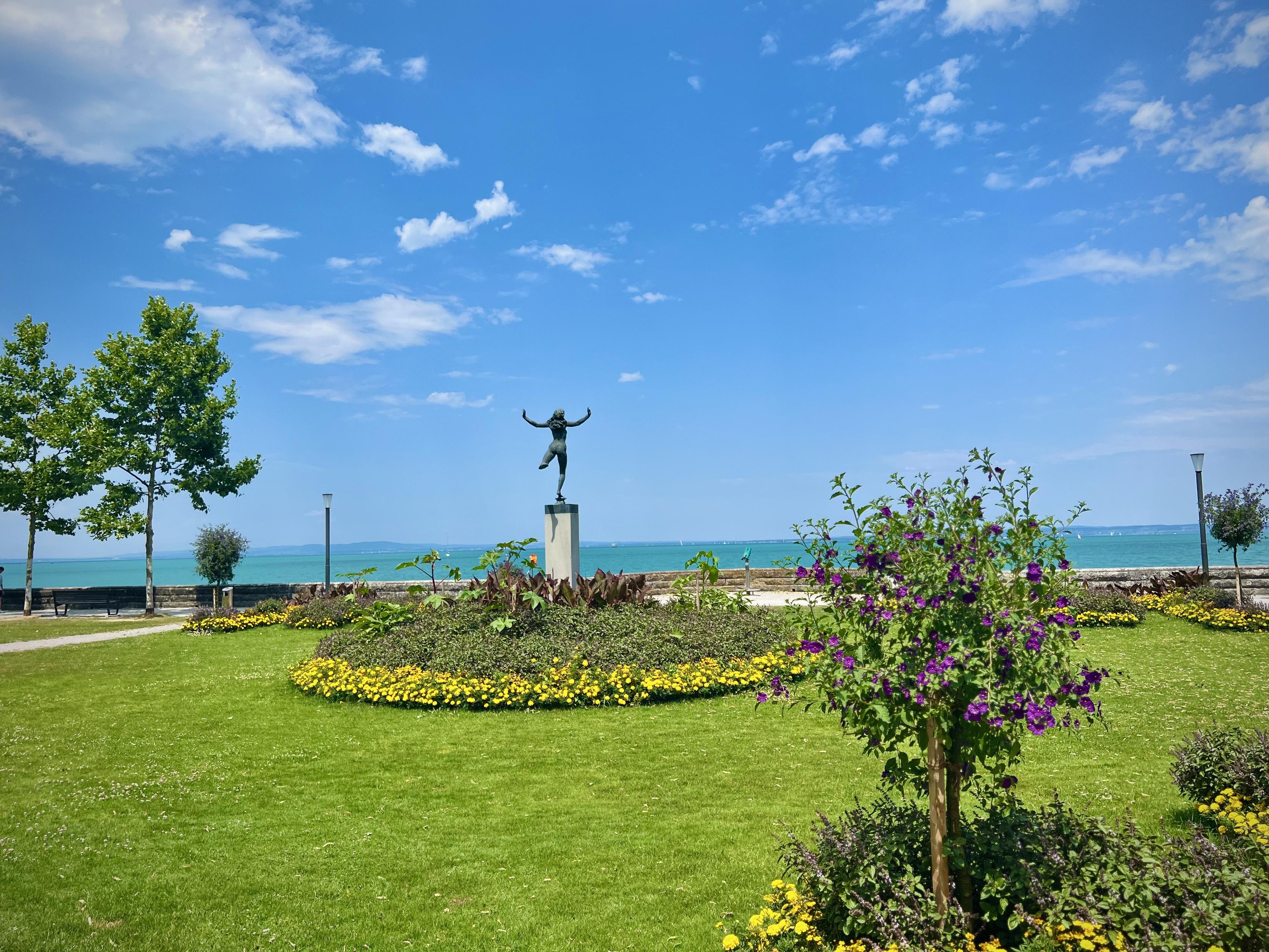 A vibrant, sunlit park by a turquoise lake. In the center of a neatly manicured green lawn, a bronze statue of a dancing or leaping figure stands atop a stone pedestal. Surrounding the base of the statue is a circular garden bed filled with small, bright yellow flowers and leafy plants.

To the right, a young shrub is covered in brilliant purple blossoms. In the background, a low stone wall separates the park from the wide, expansive lake, which stretches toward a distant, hazy shoreline. The sky is a bright, clear blue with soft, scattered white clouds, creating a peaceful and scenic atmosphere.